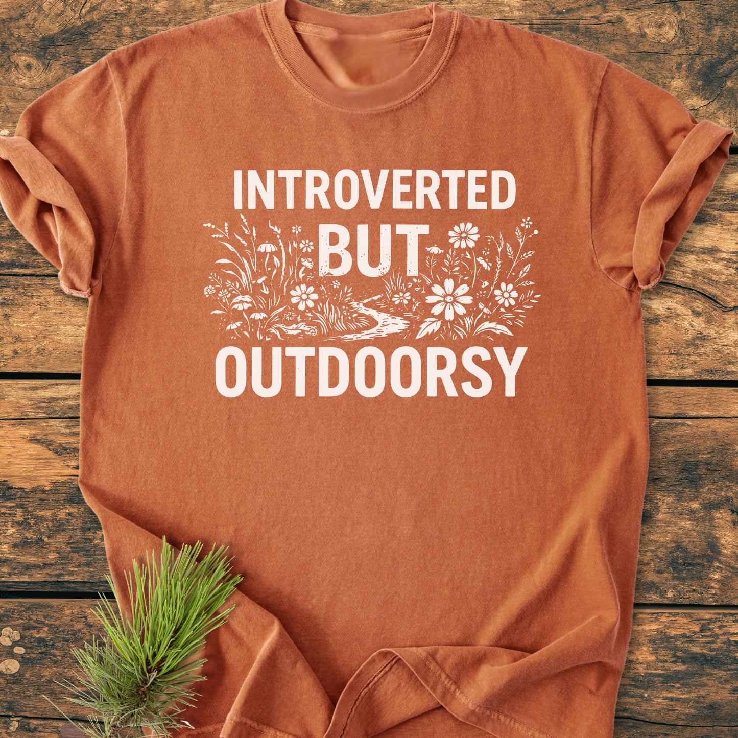 Introverted But Outdoorsy - Vintage Wash Tee: Rust-orange T-shirt with white block letters and wildflower illustration, laid on weathered wood, featuring cozy garment-dyed cotton with relaxed fit and durable stitching.