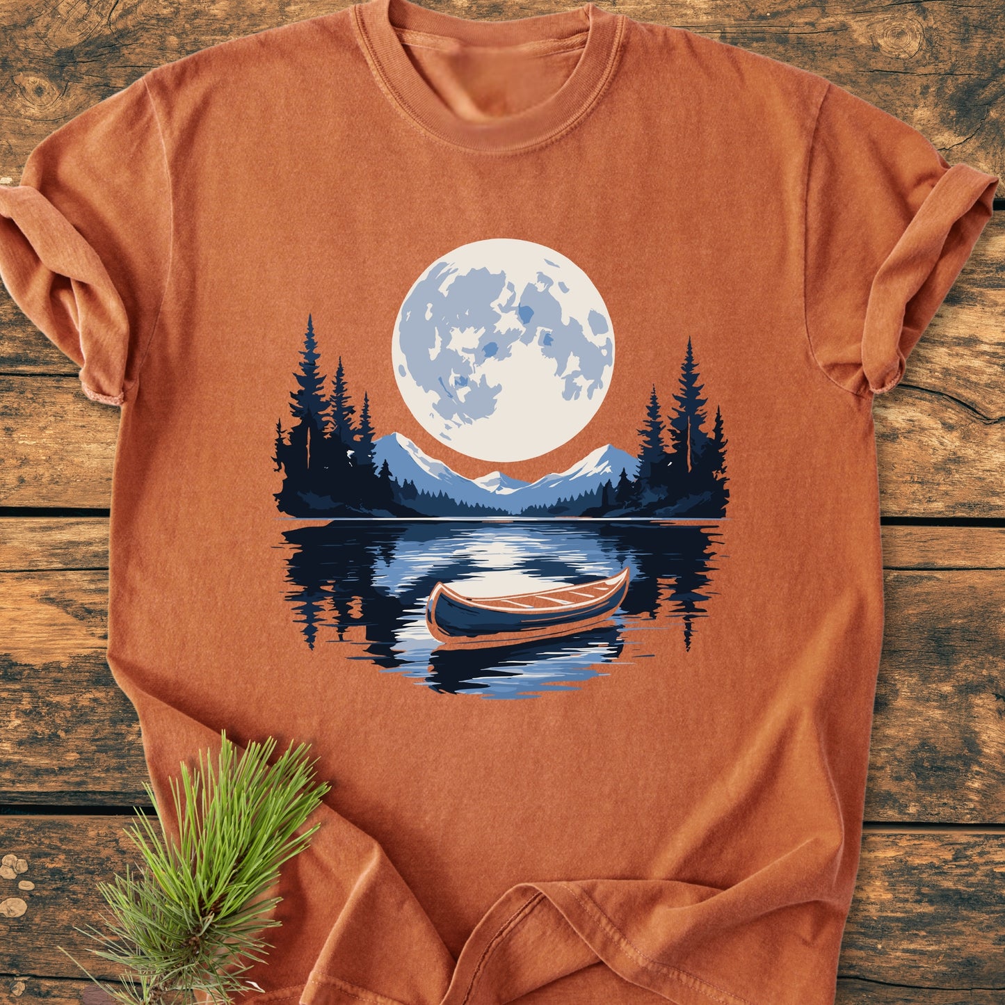 Moonlit Canoe - Vintage Wash Tee featuring a moonlit wilderness scene with mountains, trees, and a canoe on a lake, laid on a wooden surface with a pine sprig beside it.