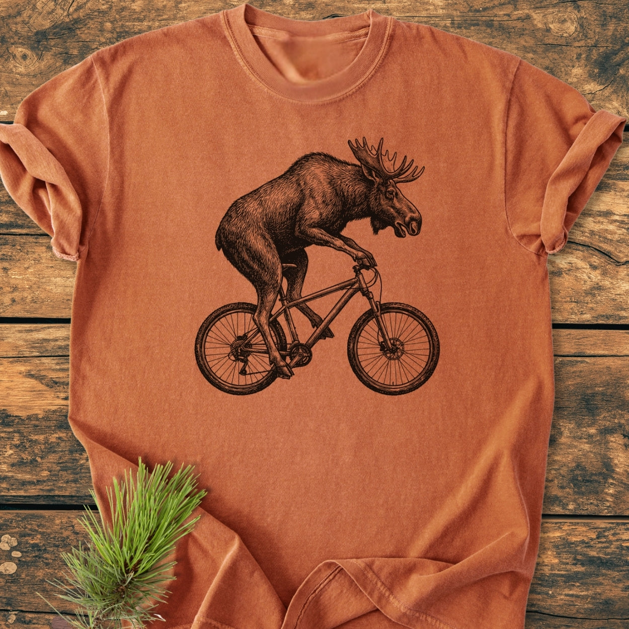 Moose On The Road - Vintage Wash Tee displays a black line drawing of a moose cycling on a heather-orange T-shirt, laid on wood, with a pine sprig beside it.