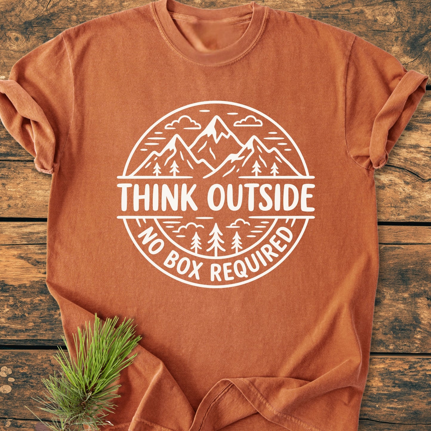 Think Outside - Vintage Wash Tee features a rust-orange hue with a white outdoorsy graphic. Laid flat on wood, it highlights comfort and durability, styled with a pine branch.