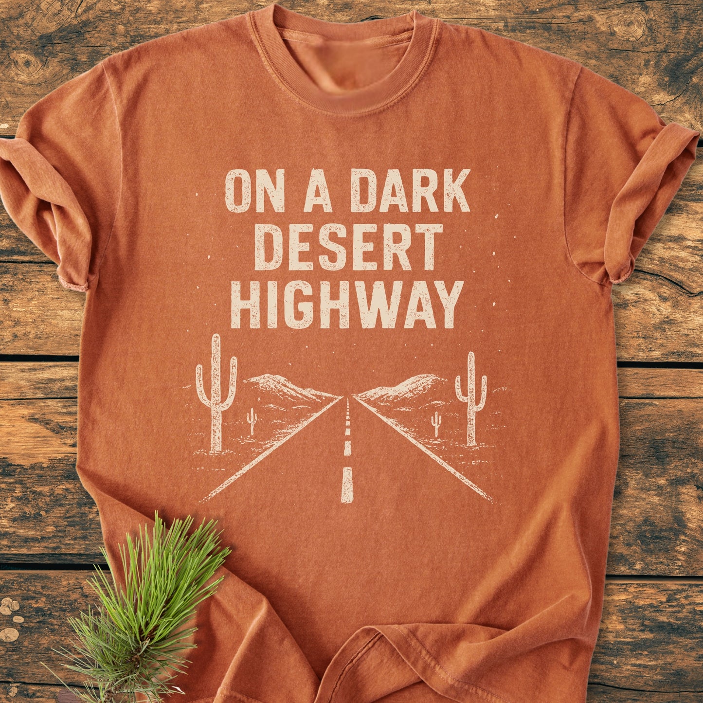 On A Dark Desert Highway - Vintage Wash Tee featuring a screen-printed road scene with cacti and mountains, laid flat on a wooden surface, paired with a pine bough for a natural touch.