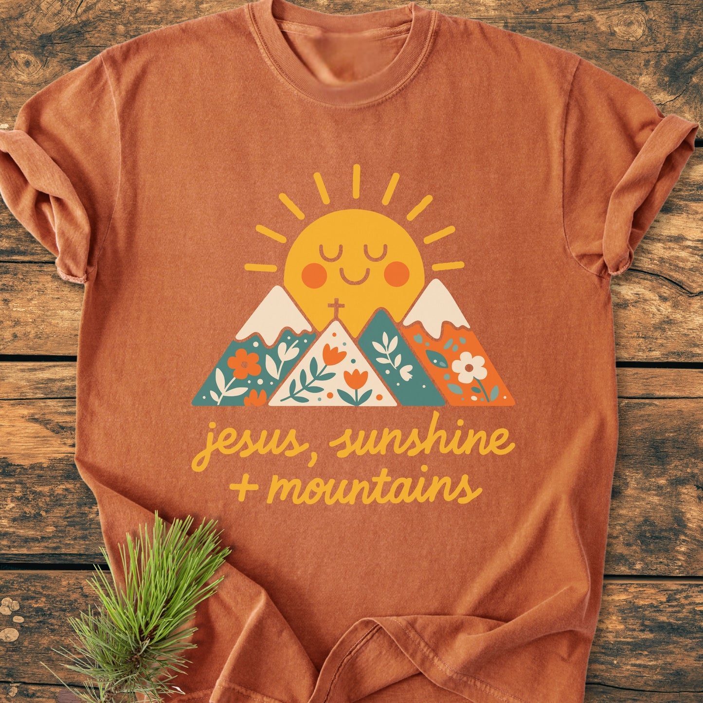 Jesus, Sunshine, Mountains - Vintage Heavyweight Tee featuring a smiling sun and floral-patterned mountains. Laid on wood, with pine sprig, highlighting the relaxed fit and garment-dyed comfort of this durable, pre-shrunk cotton tee.
