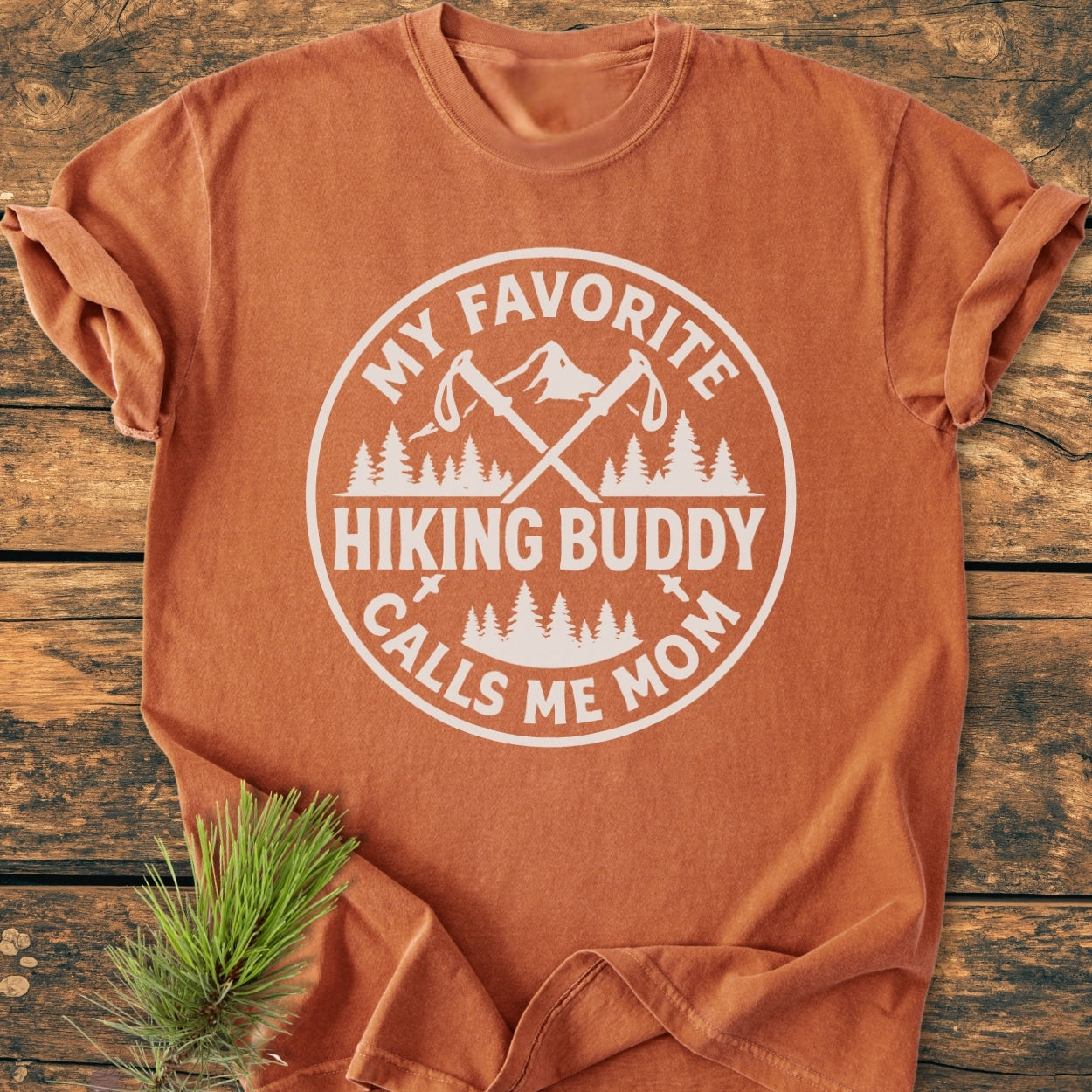The Hiking Buddy Mom - Vintage Wash Tee features a printed graphic with trekking poles and mountains, laid flat on a wooden surface. Made from soft, durable, 100% ring-spun cotton for everyday comfort.
