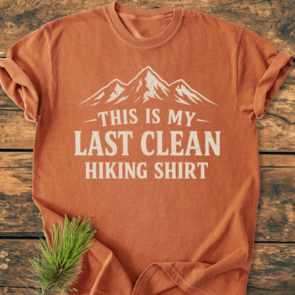 The Last Clean Hiking Shirt - Vintage Wash Tee features a rust color, mountain graphic, and THIS IS MY LAST CLEAN HIKING SHIRT text, displayed flat with a pine sprig on wood.