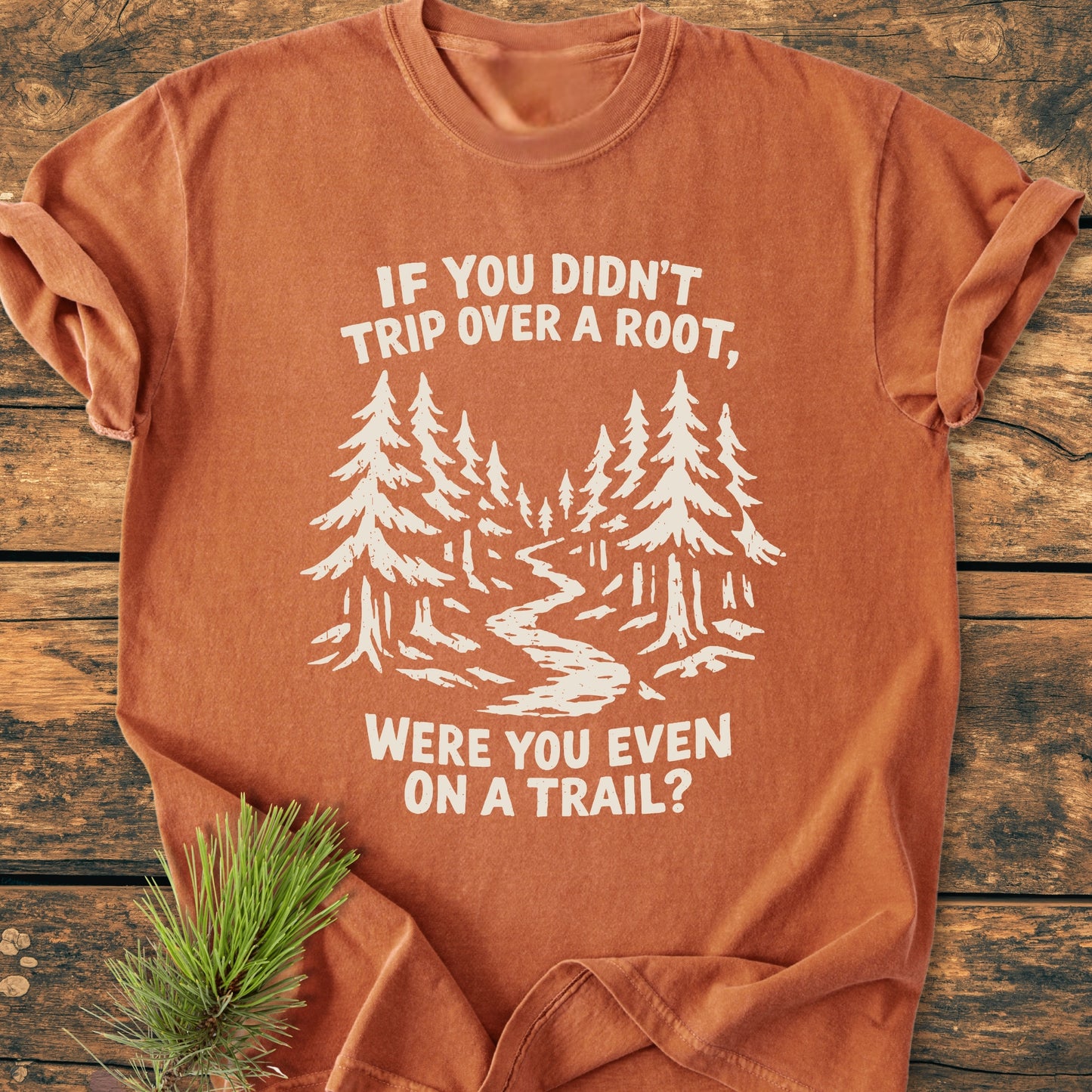 Were You Even On A Trail? - Vintage Wash Tee features a stylized forest scene with pine trees and a trail, screen-printed on a rust-orange T-shirt, laid on a weathered wood surface.