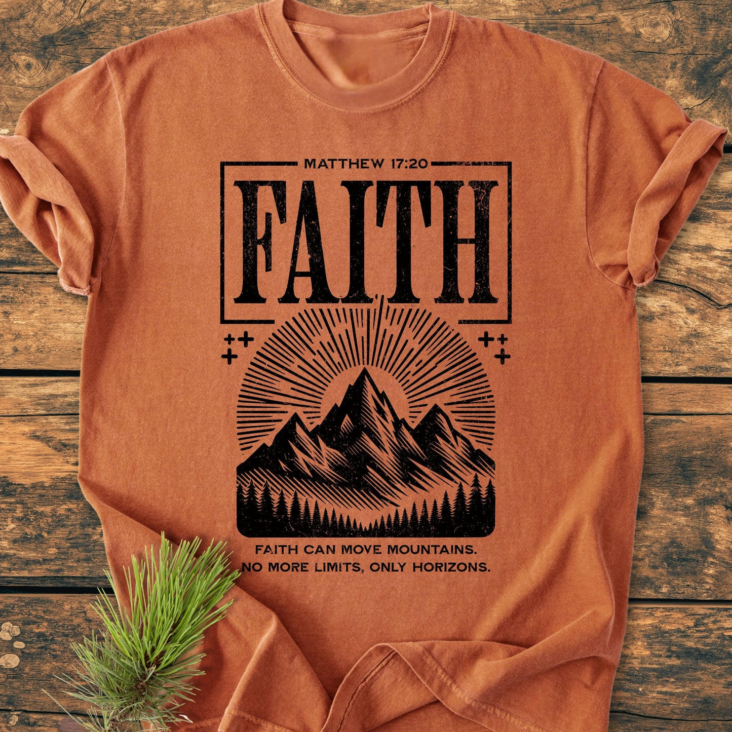 Faith Moves Mountains - Vintage Wash Tee features a black FAITH print with mountain silhouette and sunburst, laid flat on wood with a pine sprig, showcasing its relaxed fit and durable design.