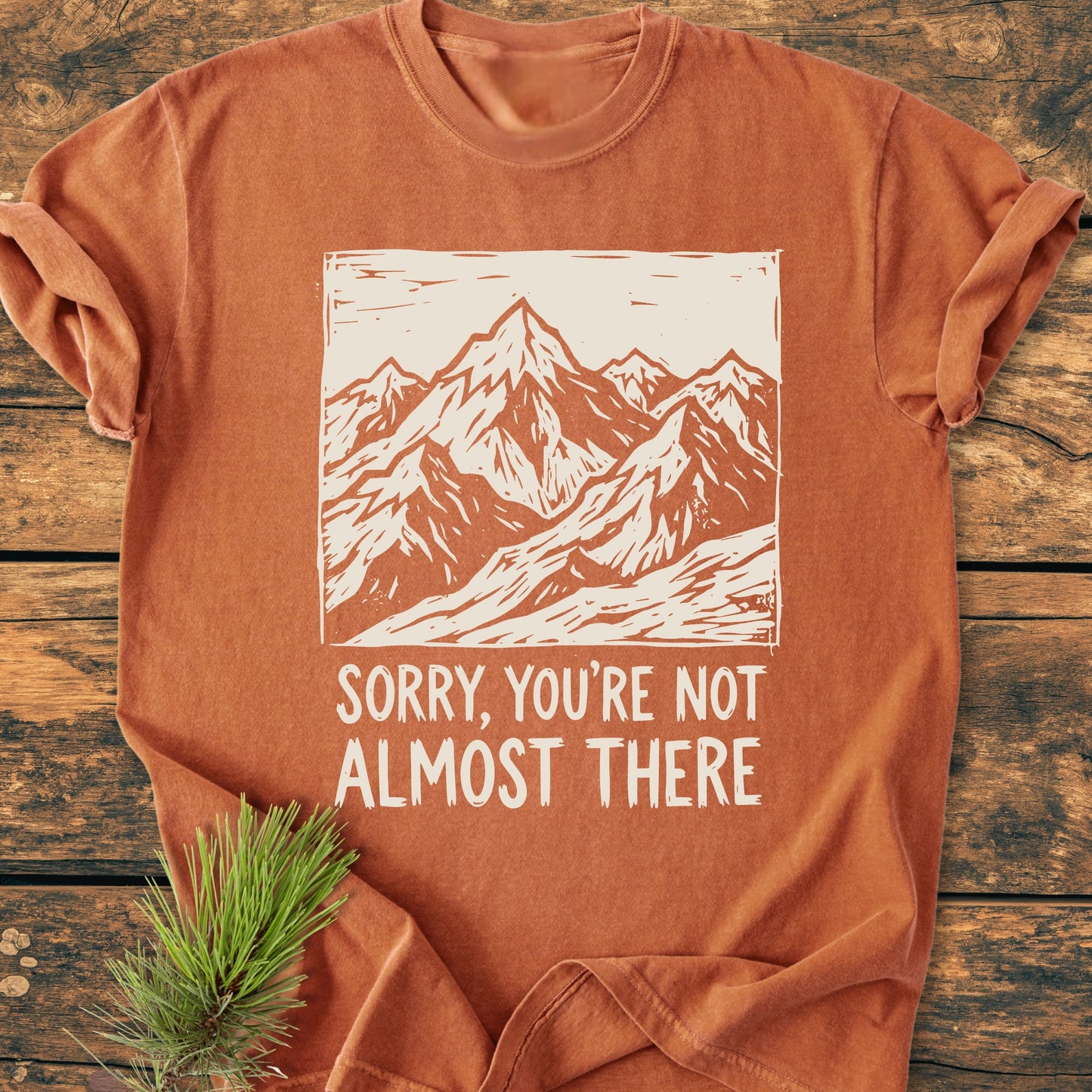 Not Almost There - Vintage Wash Tee in rust-orange, featuring a white mountain range graphic and text. Laid on wood surface with a pine branch accent, highlighting its casual, relaxed fit.