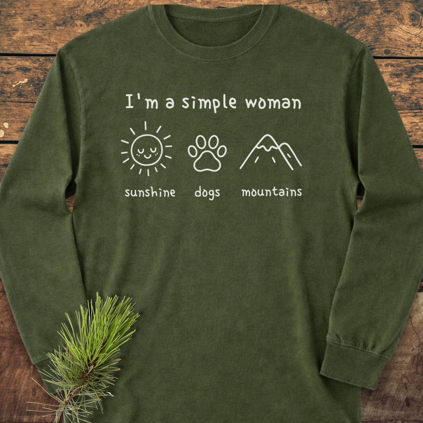 I'm A Simple Woman - Sunshine, Dogs, Mountains Long-Sleeve Tee; features forest-green fabric, chest prints of a sun, dog paw, and mountains, laid on wood with pine sprig, offering casual comfort.
