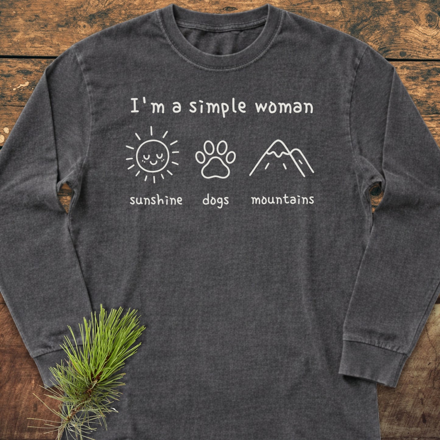 I'm A Simple Woman - Sunshine, Dogs, Mountains Long-Sleeve Tee featuring white lettering and icons on a dark-gray shirt, laid flat with a pine branch accent, emphasizing casual comfort and personalized style.