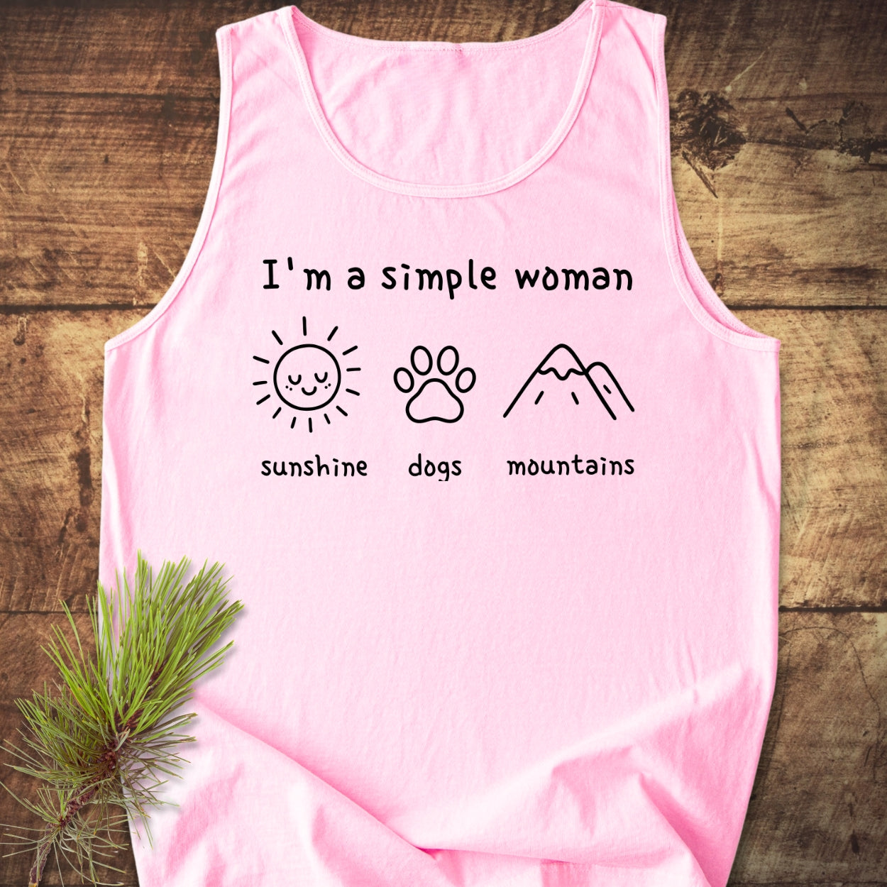 I'm A Simple Woman - Sunshine, Dogs, Mountains Tank Top features a soft pink, sleeveless design with I'm a simple woman and icons of a sun, dog paw-print, and mountains on a wooden surface.