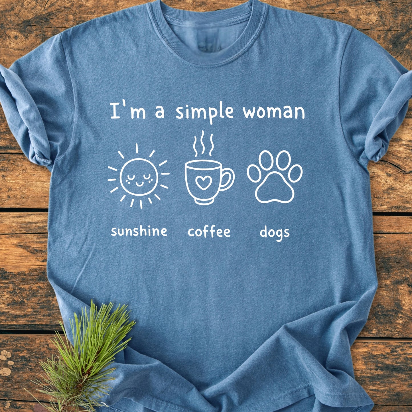 I'm A Simple Woman. Sunshine Coffee Dogs - Vintage Wash Tee, featuring three icons: sunshine, coffee, and dogs on heather-blue fabric, laid flat on rustic wood with a pine sprig.