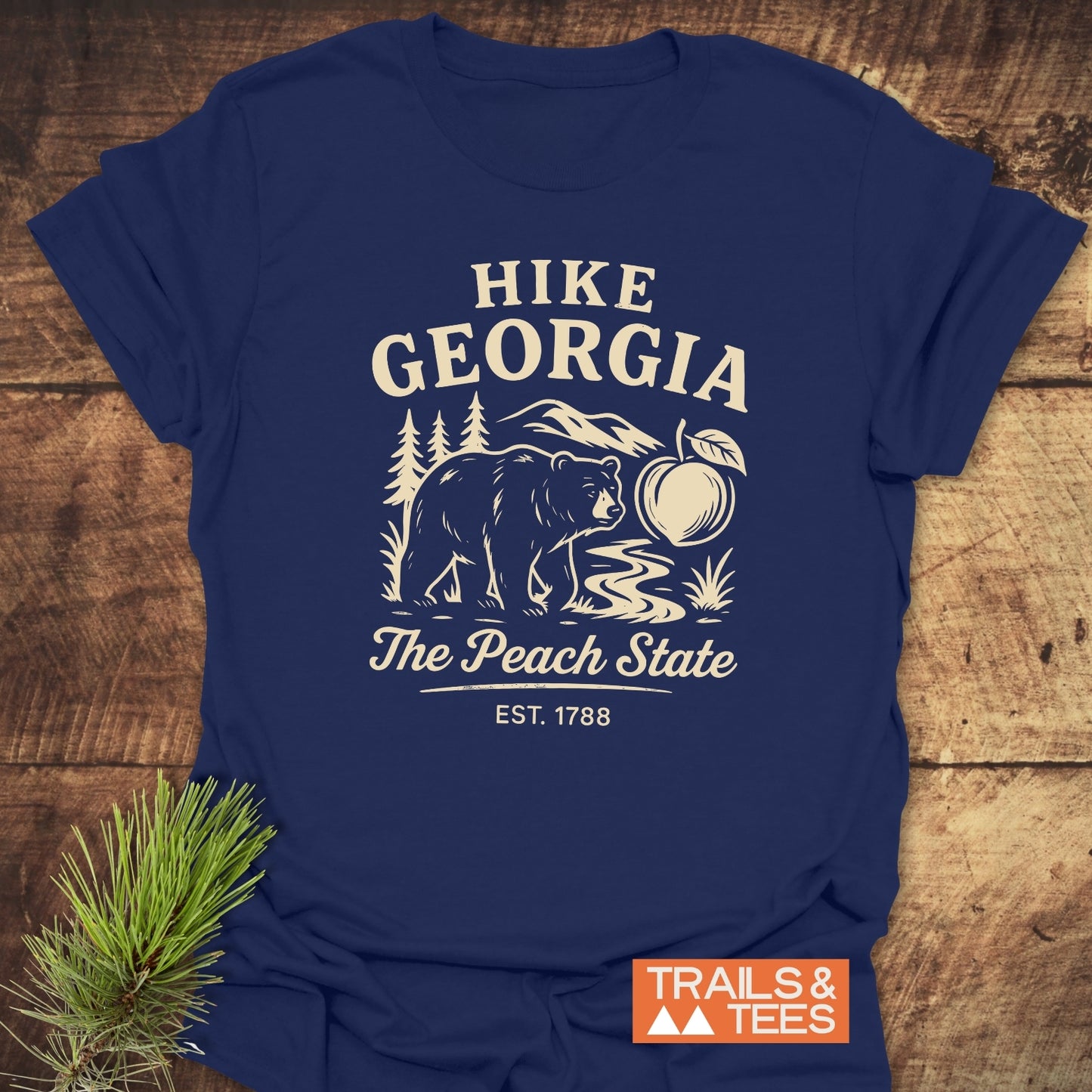 Hike Georgia T-Shirt featuring a stylized outdoor scene with mountains, trees, a bear, and a peach, framed by HIKE GEORGIA text. Includes TRAILS & TEES logo, laid on wood surface.