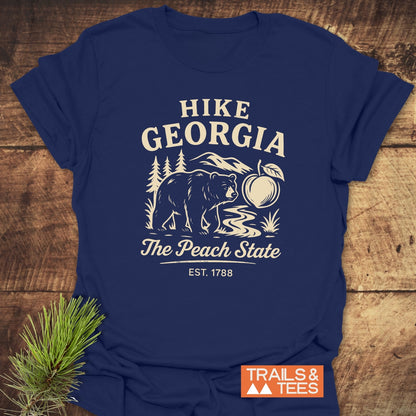 Hike Georgia T-Shirt featuring a stylized outdoor scene with mountains, trees, a bear, and a peach, framed by HIKE GEORGIA text. Includes TRAILS & TEES logo, laid on wood surface.