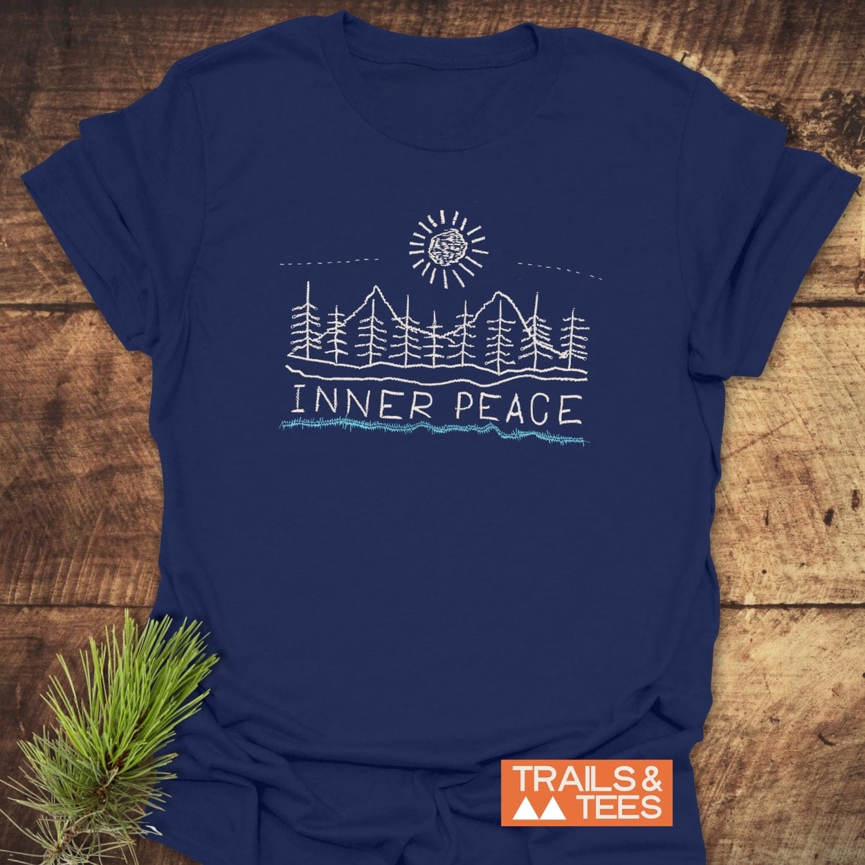 Inner Peace T-Shirt on rustic wood, featuring a white line-drawing of mountains, pine trees, and sun, with INNER PEACE text. Includes TRAILS & TEES logo and a pine branch beside it.