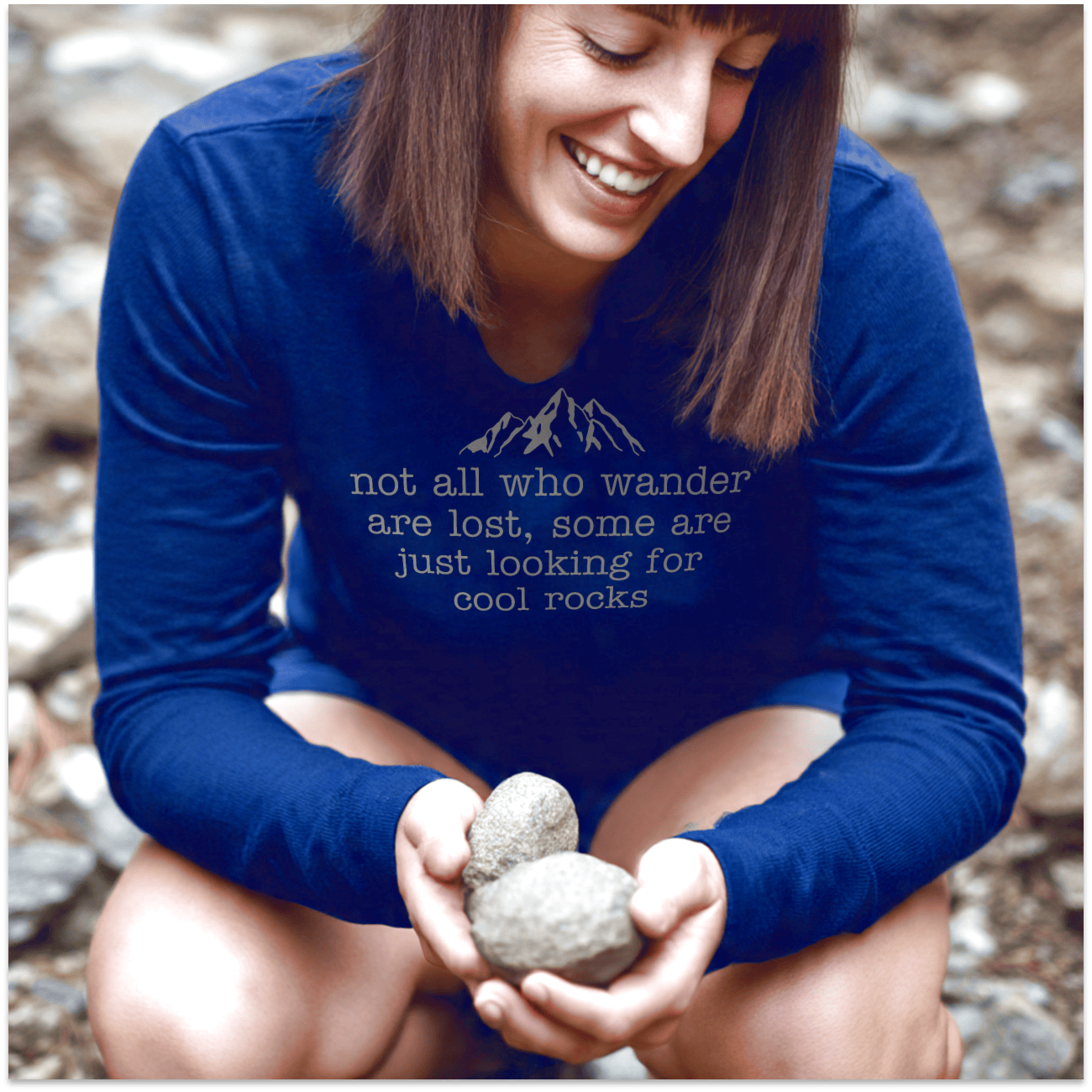Person crouched outdoors, smiling while holding stones, wearing a Looking For Cool Rocks Comfort Colors Long-Sleeve Tee, featuring mountain graphic and text.