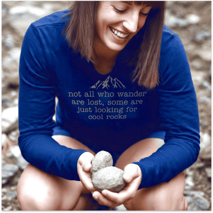 Person crouched outdoors, smiling while holding stones, wearing a Looking For Cool Rocks Comfort Colors Long-Sleeve Tee, featuring mountain graphic and text.
