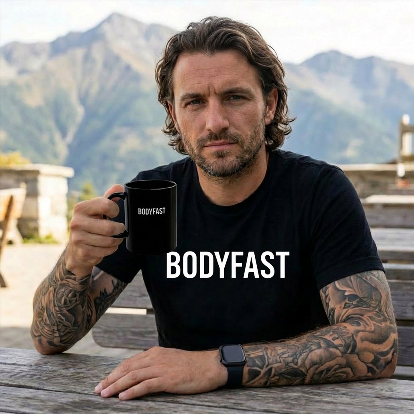 A man with tattoos wearing the BODYFAST - Vintage Wash Tee holds a matching mug, seated at a mountain-top picnic table, embodying the balanced lifestyle promoted by the BodyFast brand.