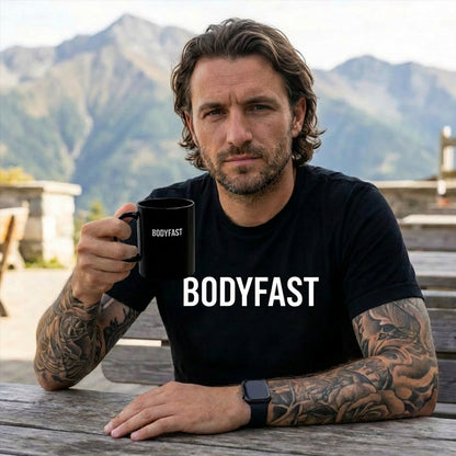 A man with tattoos wearing the BODYFAST - Vintage Wash Tee holds a matching mug, seated at a mountain-top picnic table, embodying the balanced lifestyle promoted by the BodyFast brand.