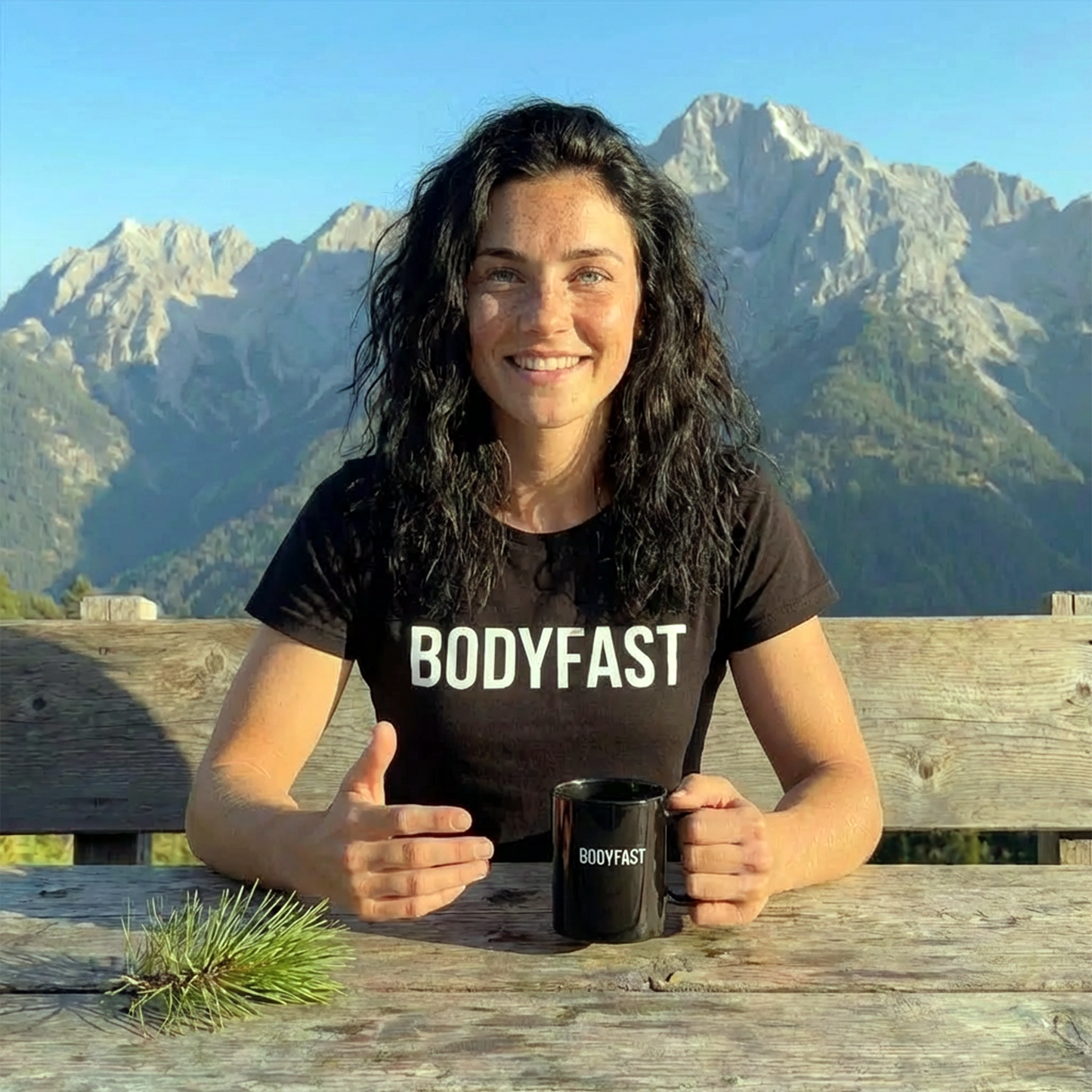 Young woman wearing a BODYFAST - Softstyle T-Shirt smiles, holding a matching mug at a picnic table, with sunlit mountains in the background, embodying a lifestyle of energy and balance.