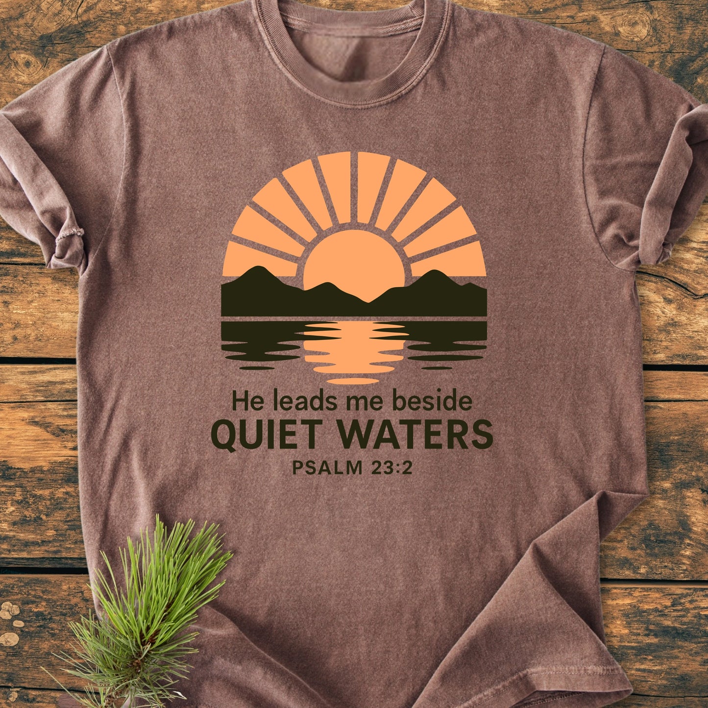 Brown T-shirt, 1424 He Leads Me, features a sunrise over mountains, text He leads me beside QUIET WATERS, Psalm 23:2, laid on wood with pine sprig, vintage soft-wash.