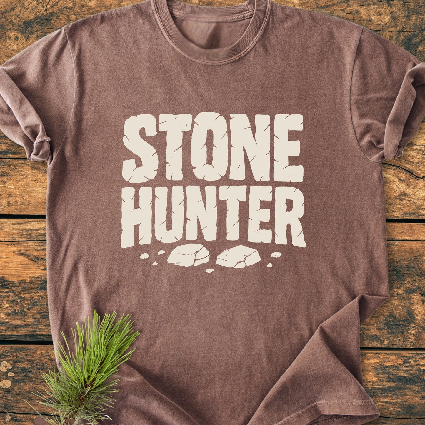 Brown/tan crew-neck T-shirt, STONE HUNTER graphic in cracked-stone style, laid flat on a wooden surface. Features a vintage wash for a soft, broken-in feel, ideal for everyday wear.