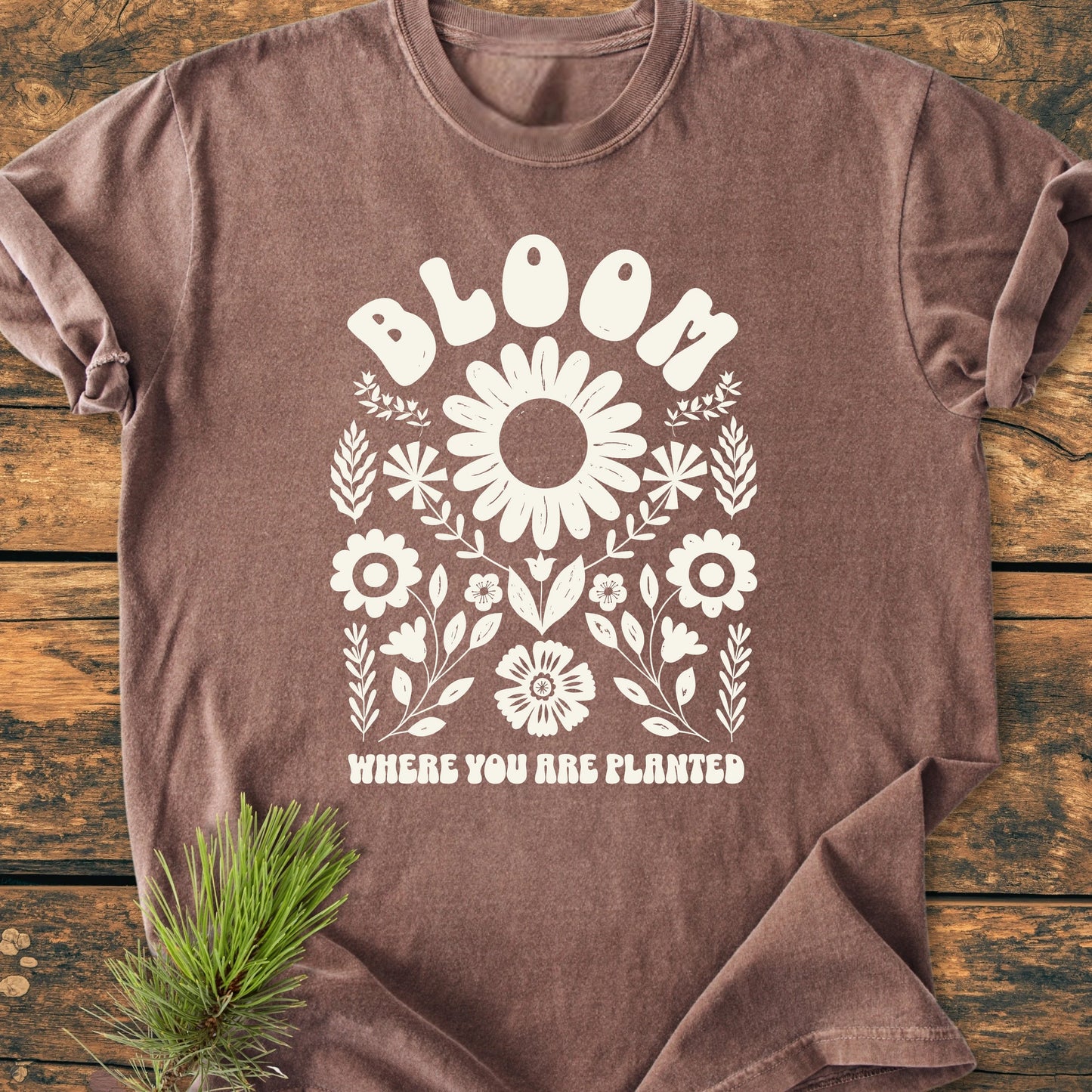 Folded 1415 Bloom Where You Are Planted T-shirt with a white floral design and phrase, laid on wood with a pine branch. Vintage wash, relaxed fit, soft cotton.