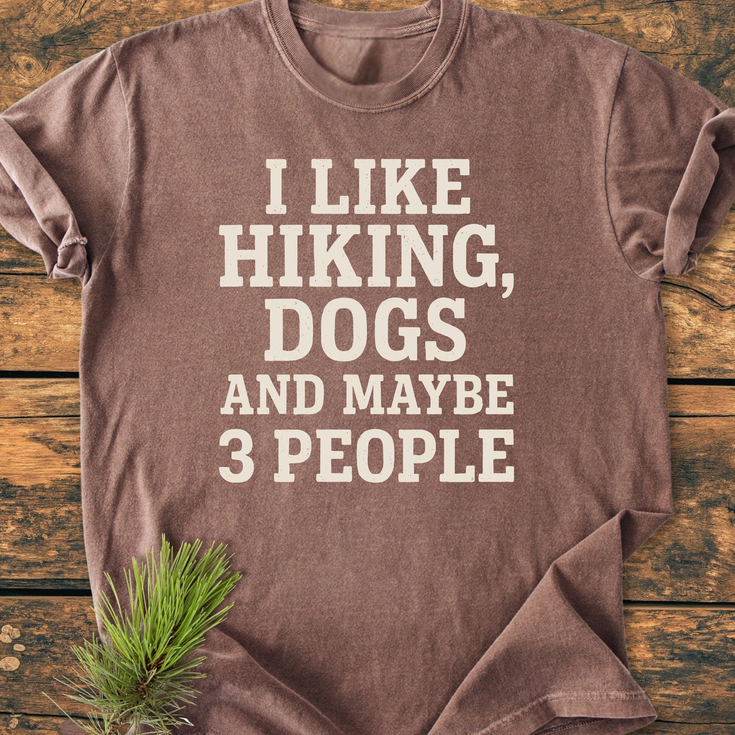 Brown-heather T-shirt with I LIKE HIKING, DOGS AND MAYBE 3 PEOPLE in off-white block letters, laid on wood, pine sprig nearby. Vintage wash, relaxed fit, ideal for outdoor enthusiasts.