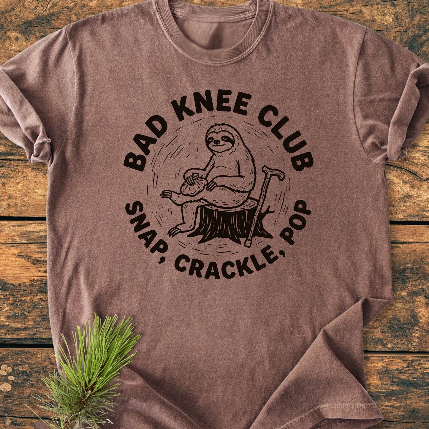 Brown T-shirt displaying Bad Knee Club with a cartoon sloth on a tree-stump, holding an ice-pack, and cane beside. Below, it reads Snap, Crackle, Pop. Laid on wooden surface.