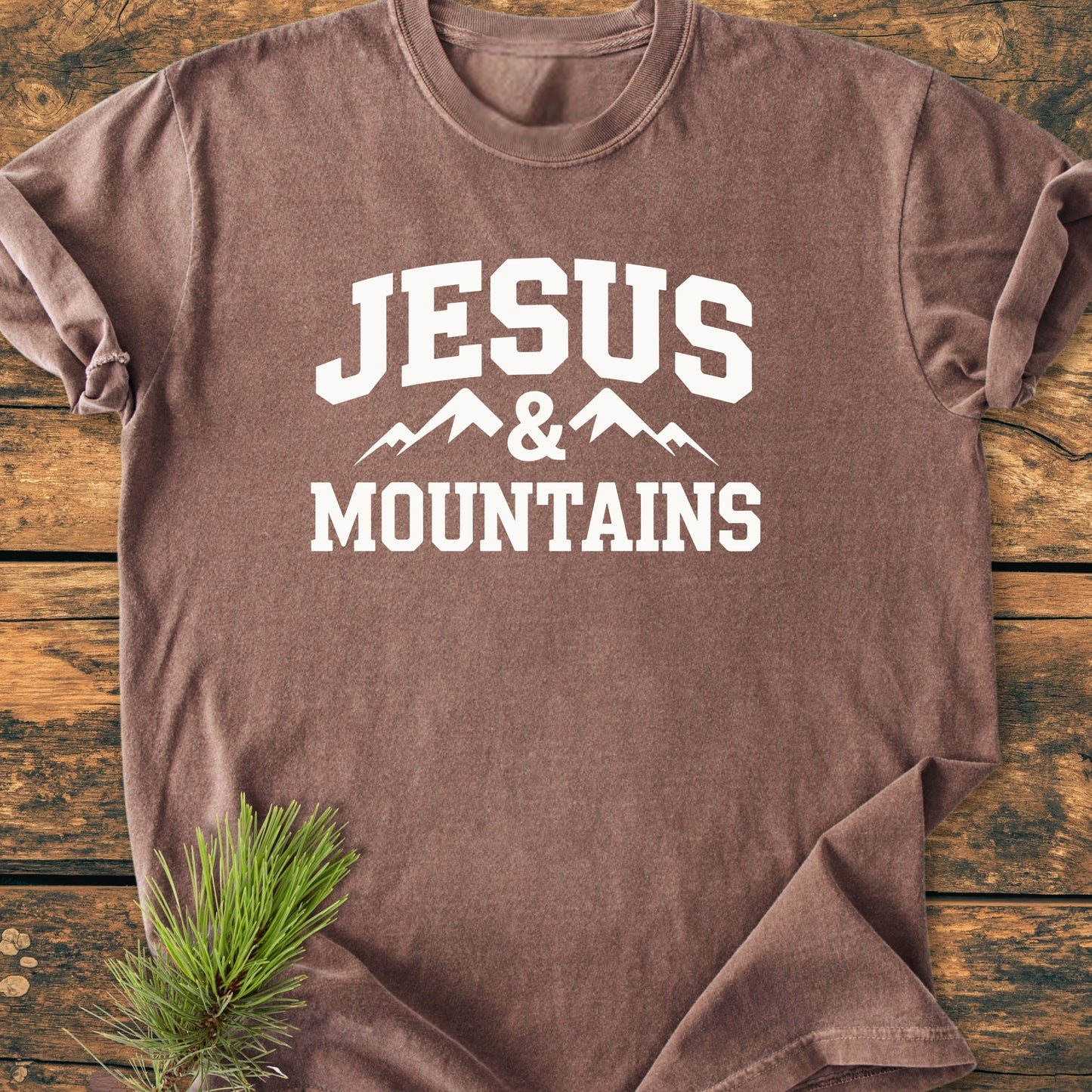 Brown crew-neck T-shirt with JESUS & MOUNTAINS and mountain graphic, laid flat on wood. Designed for comfort and durability, it embodies a vintage wash and relaxed fit.