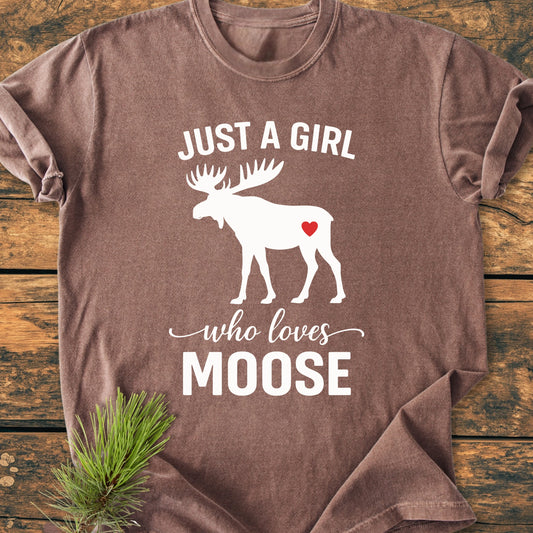 Brown T-shirt with 1437 Just A Girl Who Loves Moose text and moose silhouette, laid on wood, conveying a cozy, vintage-wash design ideal for nature enthusiasts.