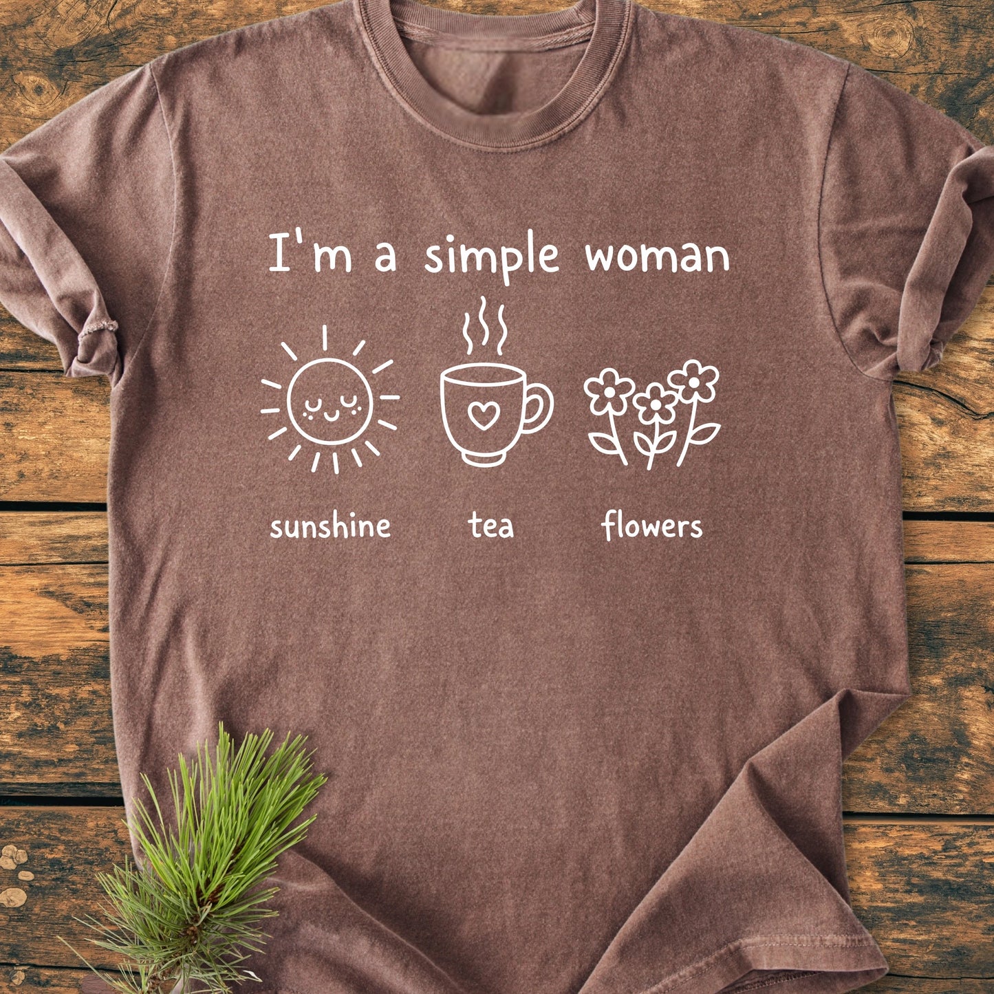 Brown 1408 I'm A Simple Woman T-shirt features white graphics with sunshine, tea, and flowers on a rustic wooden surface, with a pine sprig tucked under one edge.