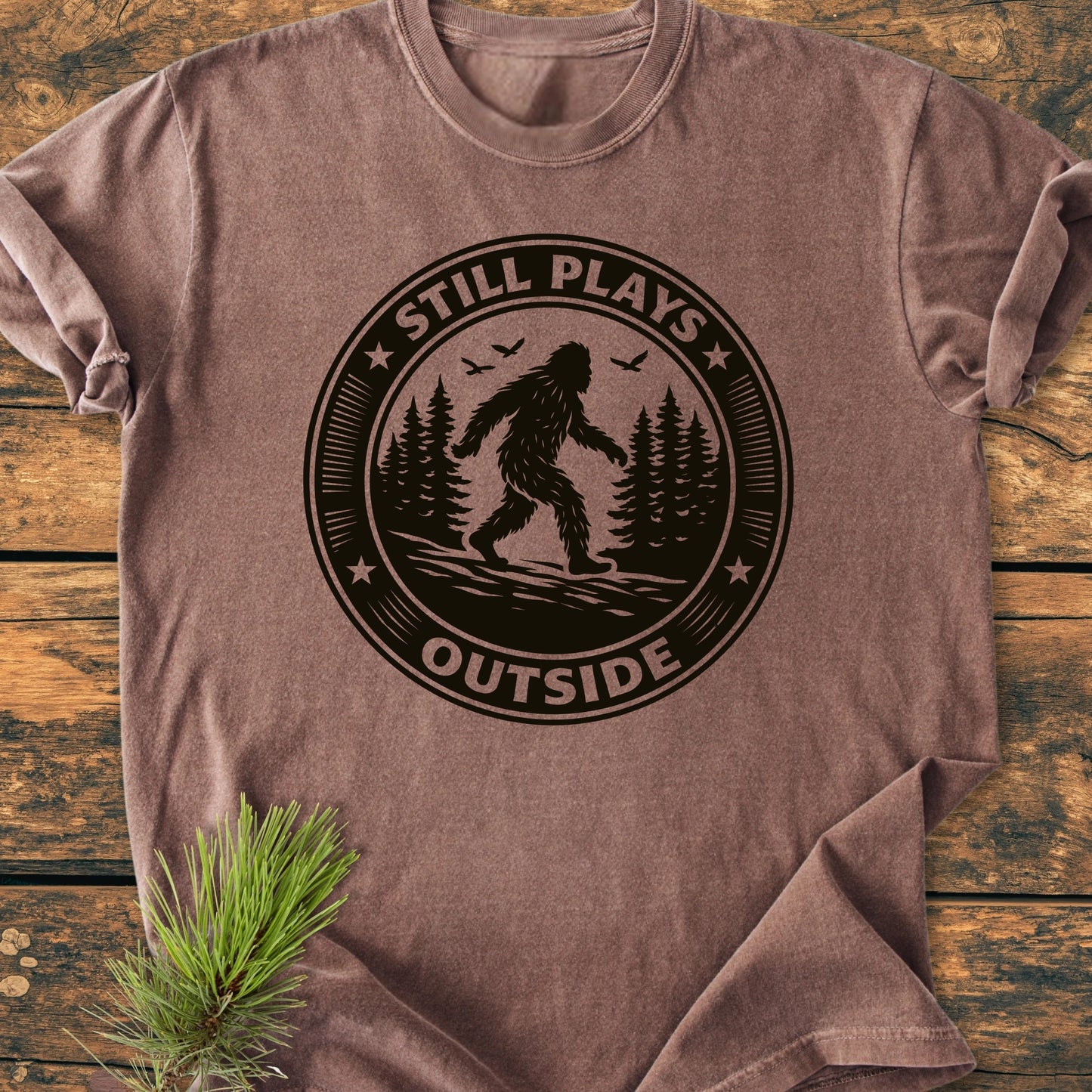 Brown T-shirt with 1430 Still Plays Outside Bigfoot graphic, featuring Bigfoot in a pine forest. Laid on wood with a pine sprig, showcasing vintage wash and relaxed fit.