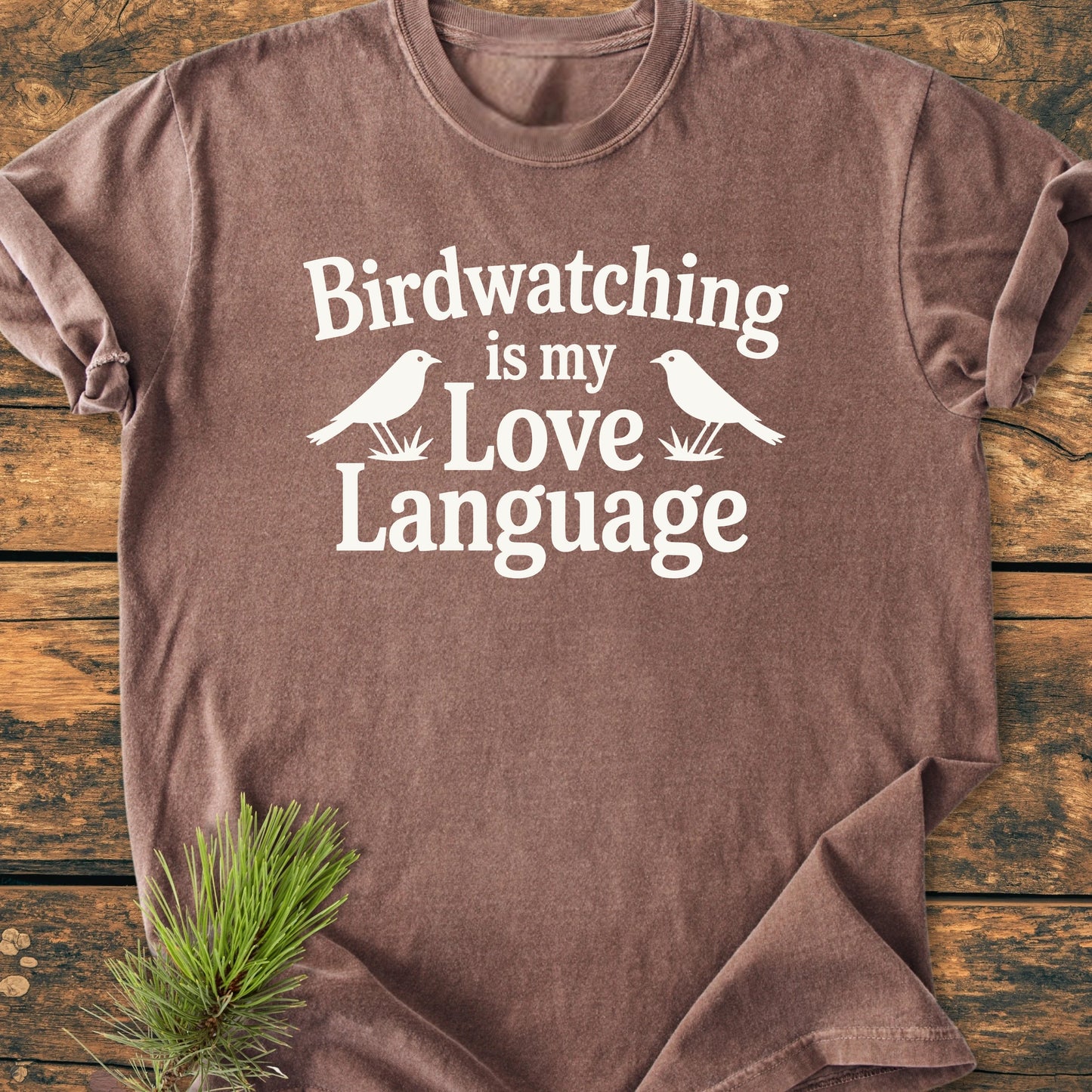 Heathered brown tee, 1428 Birdwatching Love Language, with white text and bird silhouettes, laid on wood. Styled with pine branch. Soft, vintage wash, relaxed unisex fit, durable stitching.