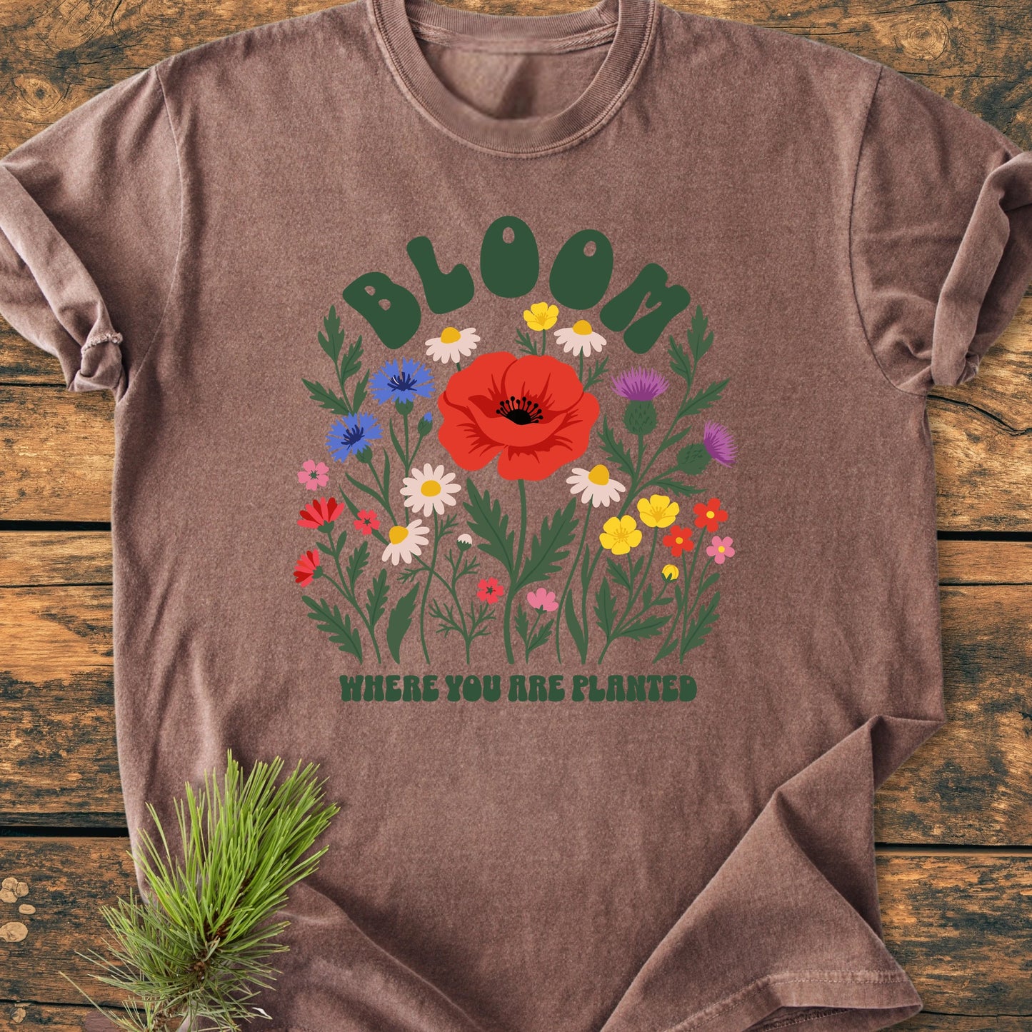 Heather-brown 1403 Bloom Where You Are Planted Base T-shirt with colorful wildflower graphic and BLOOM text, laid on wood, pine sprig beside it. Relaxed fit, 100% ring-spun cotton.