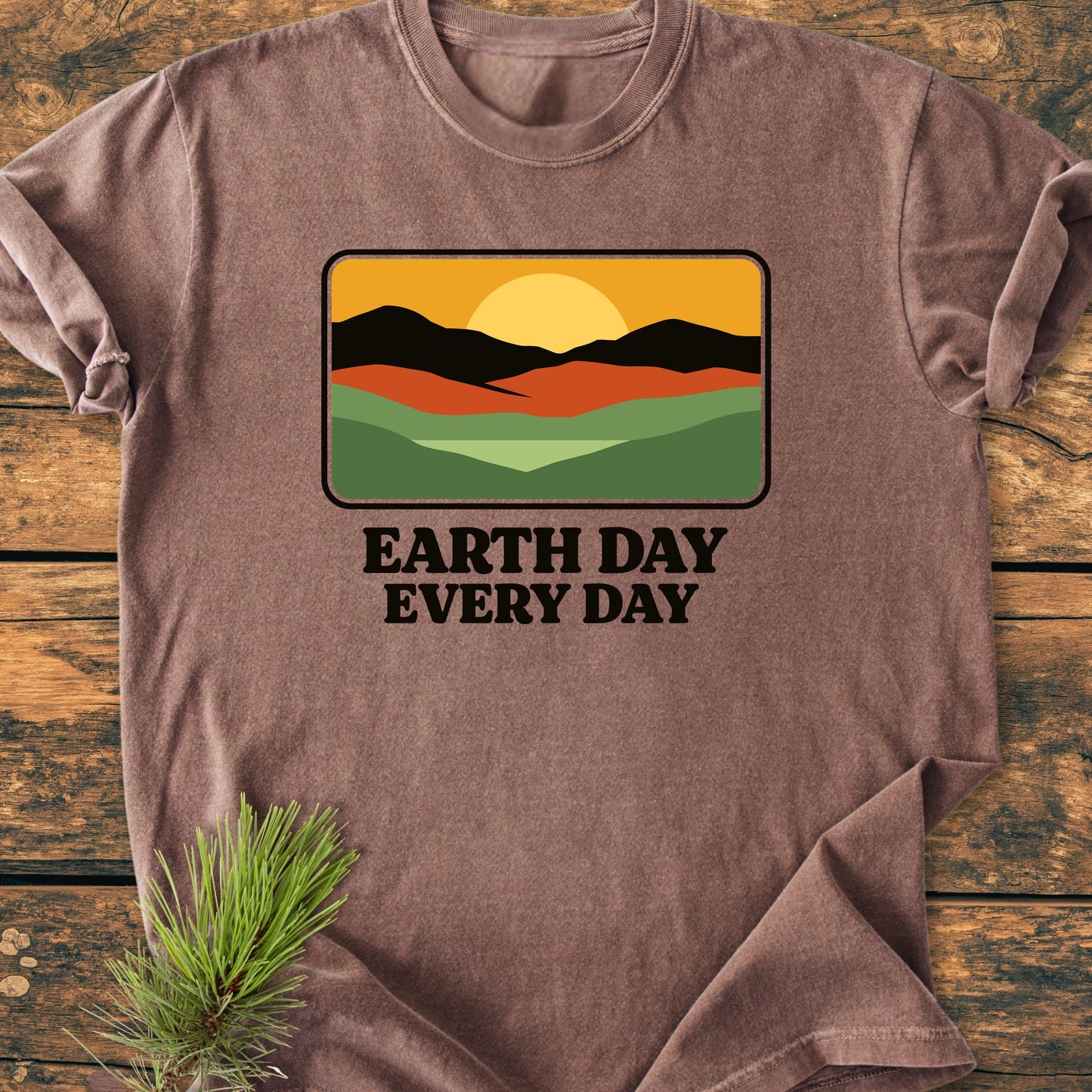 Heather-brown T-shirt featuring a retro graphic of hills, mountains, and sun, with EARTH DAY EVERY DAY text. Laid flat on wood surface, emphasizing its vintage wash and relaxed fit.