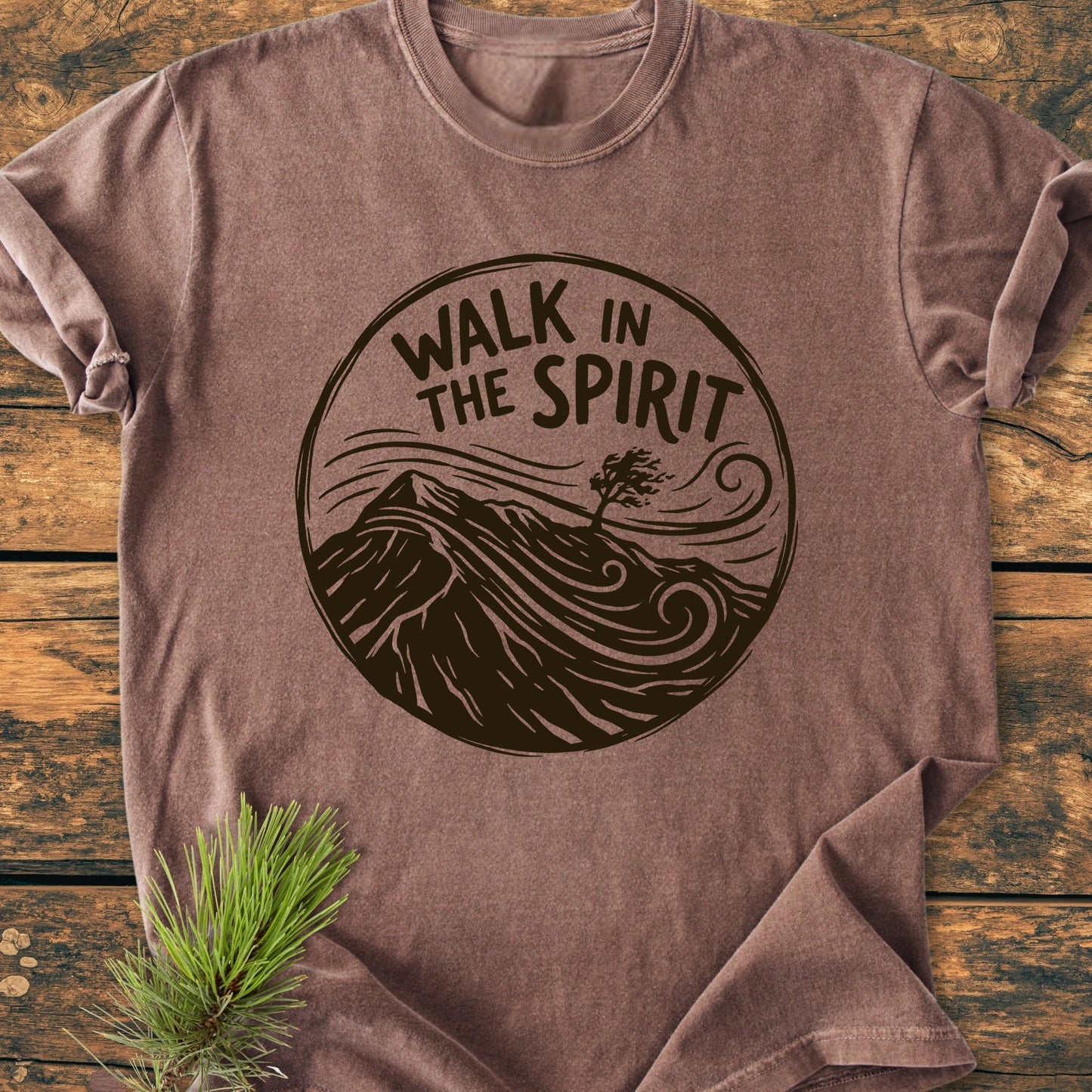 Brown cotton T-shirt, 1410 Walk In The Spirit, features a dark-brown mountain graphic with swirling winds. Laid flat on wood, showcasing a relaxed fit and durable stitching, perfect for everyday comfort.
