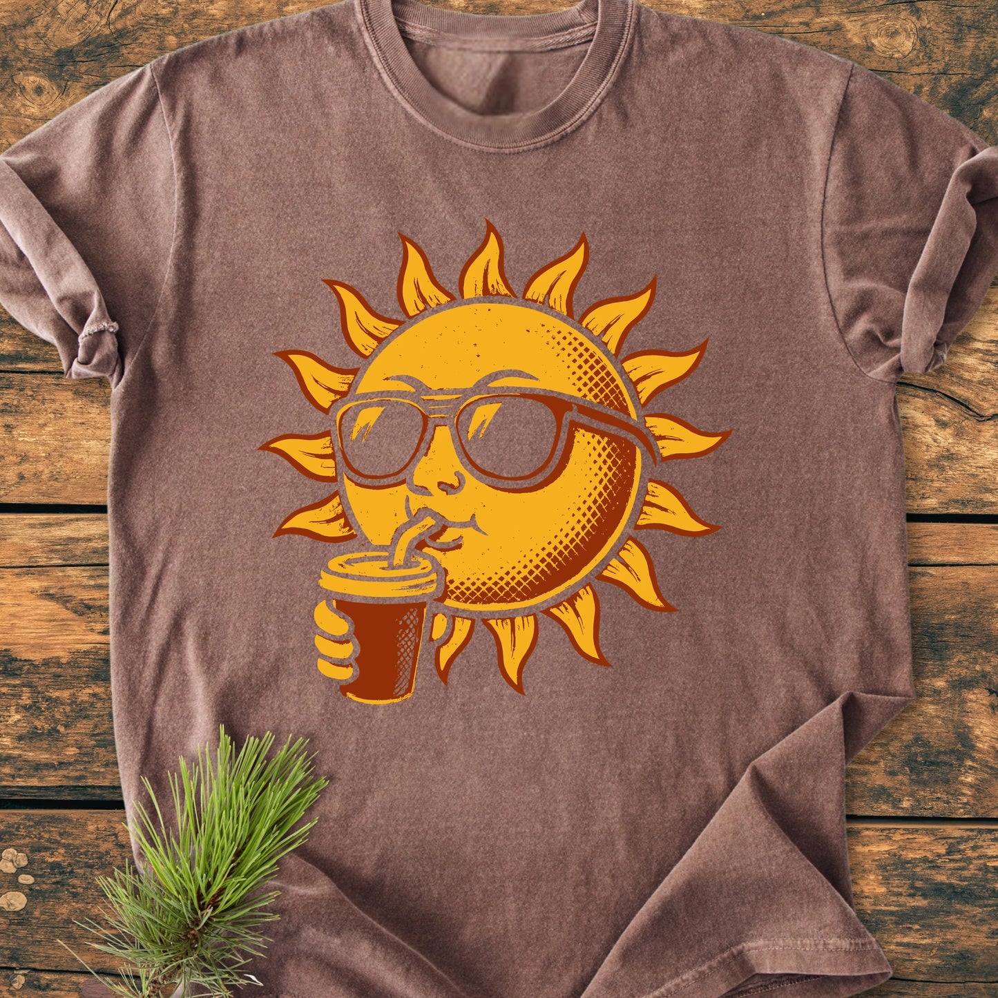 235 Sun With Coffee T-shirt features a playful sun graphic sipping from a cup. Laid flat on a wooden surface, showcasing its relaxed fit and durable, garment-dyed 100% ring-spun cotton fabric.