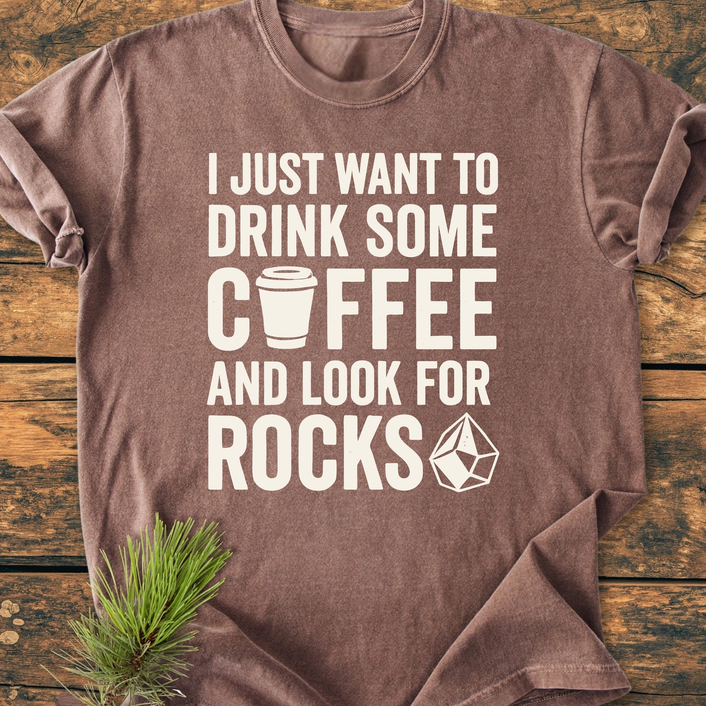 Brown short-sleeved 1404 Drink Coffee And Look For Rocks T-shirt featuring white text with coffee cup and crystal icons, laid flat; decorated with a pine sprig, highlighting relaxed fit and durable construction.
