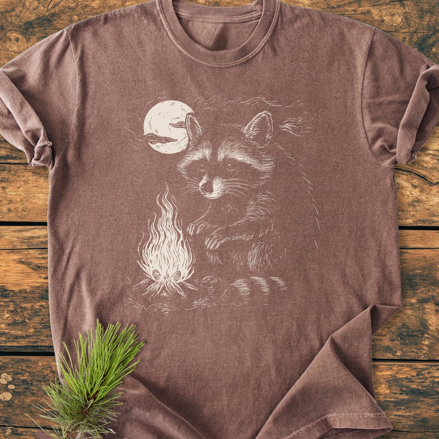 The 1409 Moonlit Campfire Raccoon Base t-shirt features a raccoon by a campfire under a full moon, screen-printed on a soft brown tee, laid flat on a wooden surface.