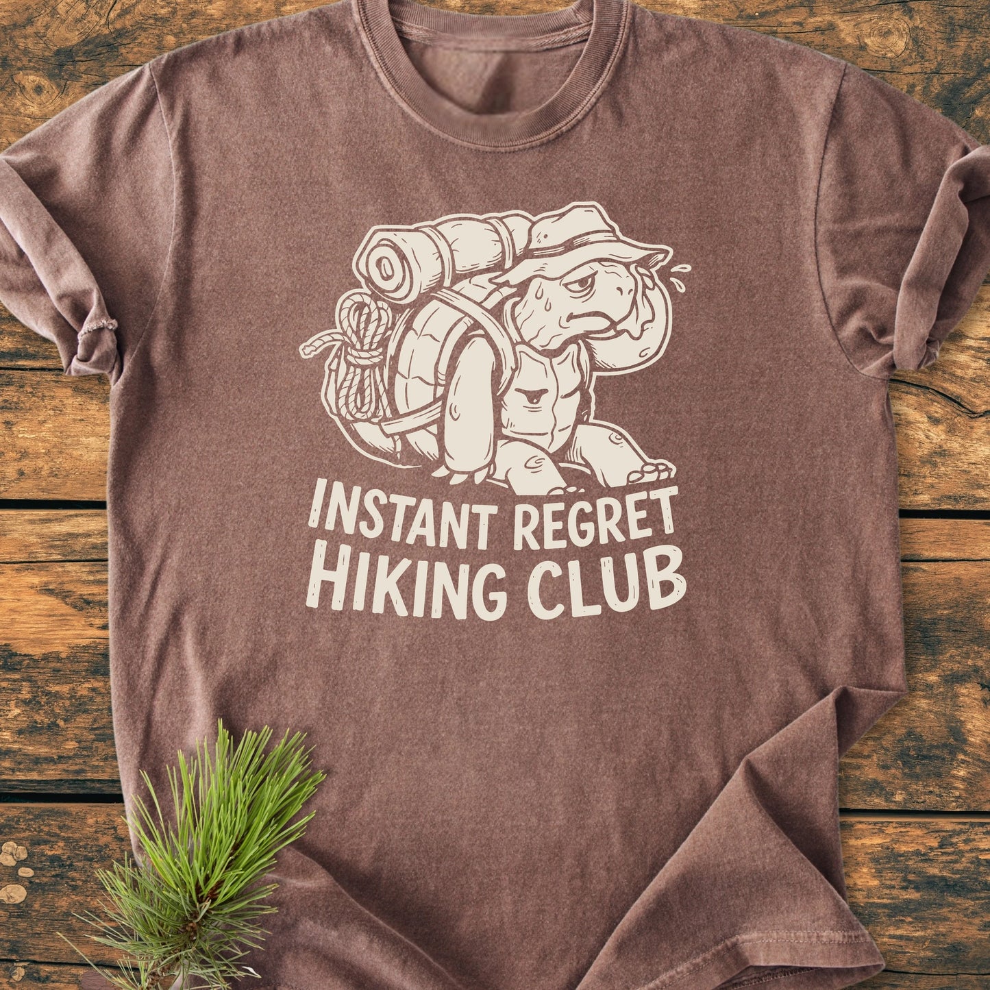 Brown T-shirt featuring 1430 Instant Regret Hiking Club slogan with cartoon tortoise in a bucket hat carrying an overloaded pack, styled with a pine sprig on a wooden surface.