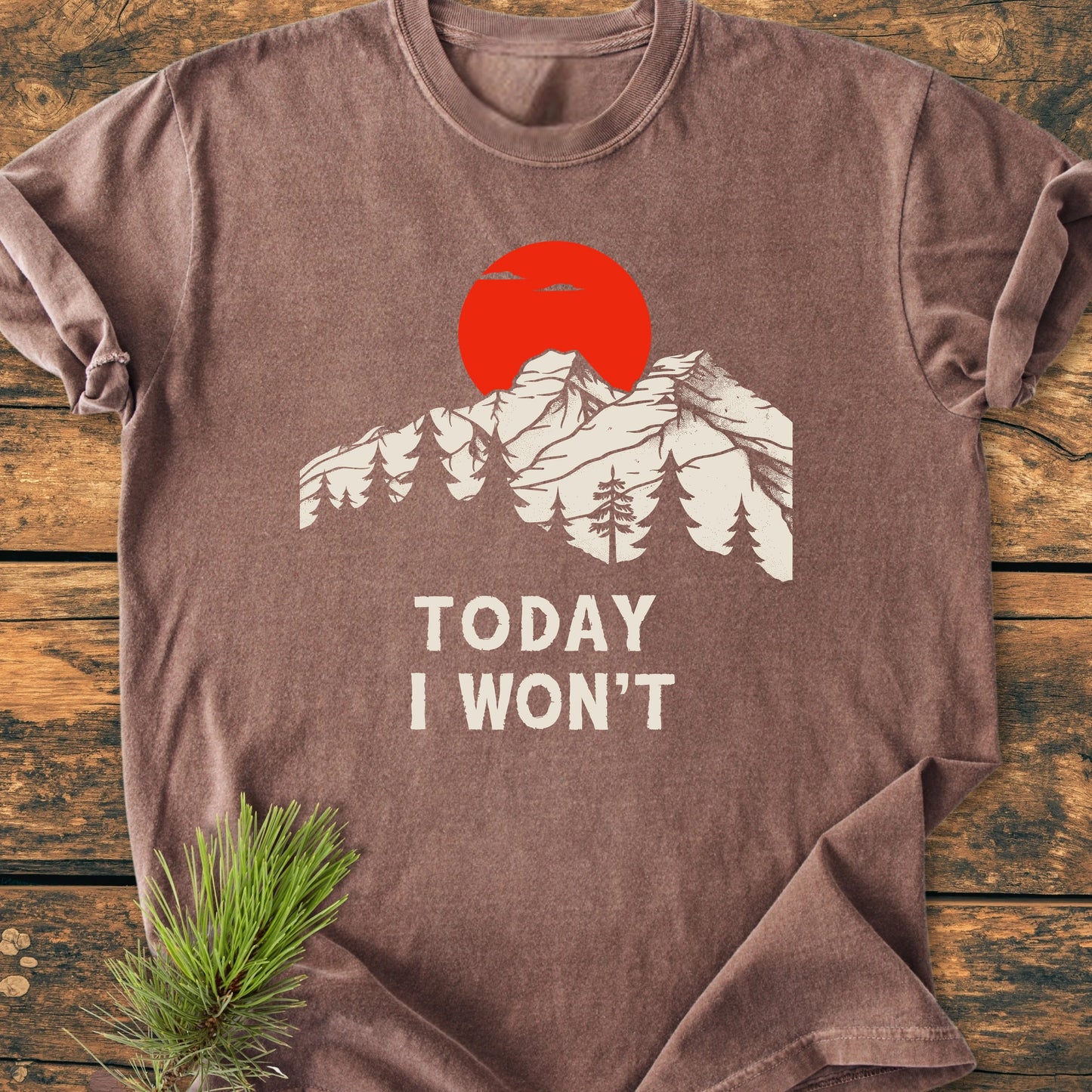 Brown crew-neck T-shirt 1423 Today I Won't with a vintage mountain-and-forest graphic, red sun, and slogan. Laid on wood with pine branch, reflecting a relaxed, vintage-wash style.