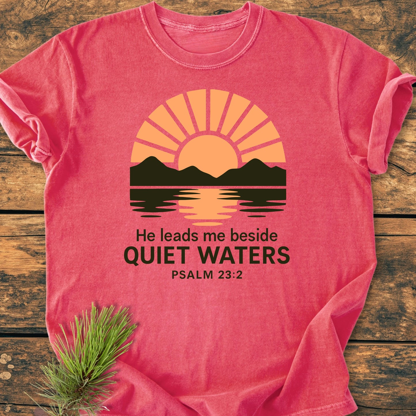 Coral-pink T-shirt, 1424 He Leads Me, features a sunlit mountain graphic and Psalm 23:2 text, laid on wood with pine sprig, embodying a soft, vintage wash for everyday comfort.