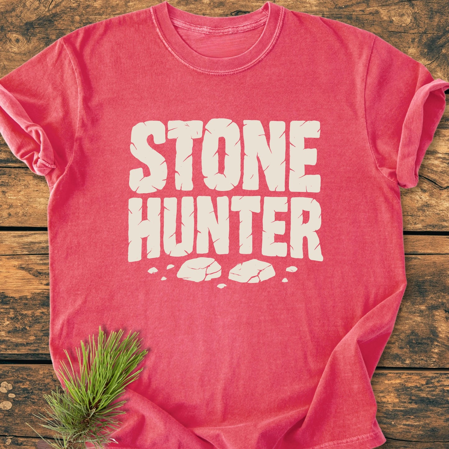 Pink-red vintage wash tee with STONE HUNTER graphic and rock fragments, laid flat on wood. Features garment-dyed, relaxed fit, highlighting Trails & Tees' durable, lived-in design. Pine sprig accent.