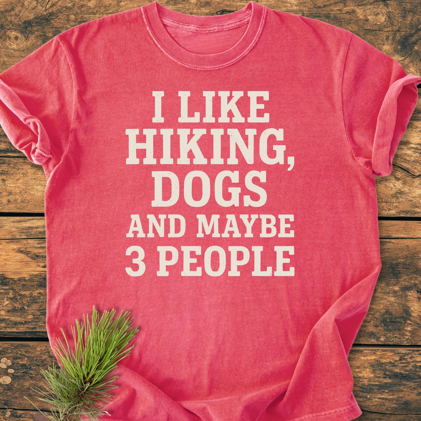 Coral-red 1431 I Like Hiking And Dogs T-shirt on rustic wood, featuring bold off-white slogan text. Accompanied by a pine sprig, showcasing vintage wash and relaxed unisex fit.