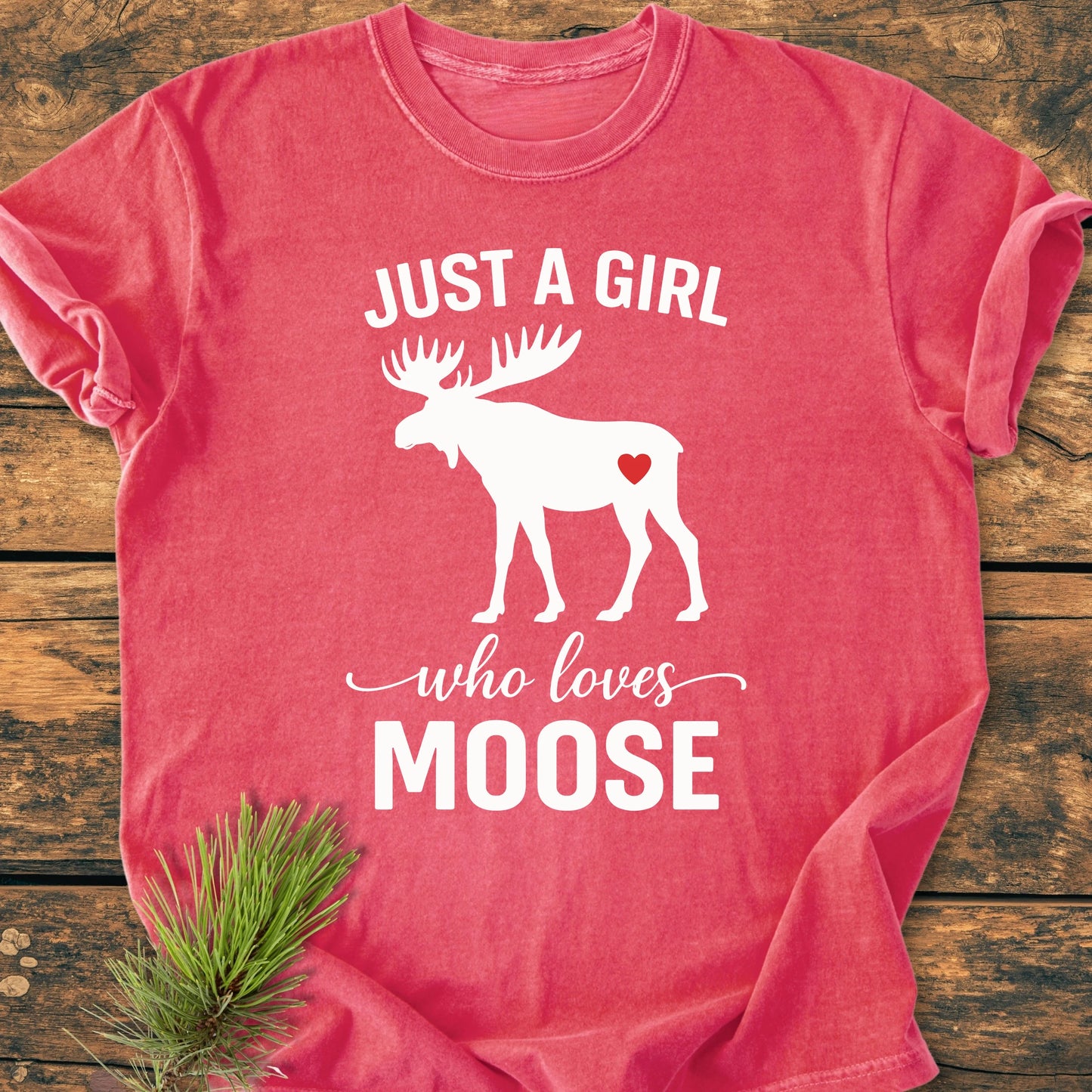 Red-pink T-shirt with white moose silhouette and JUST A GIRL who loves MOOSE slogan, featuring a red heart, laid on wood with pine sprig; vintage wash, soft, unisex fit.