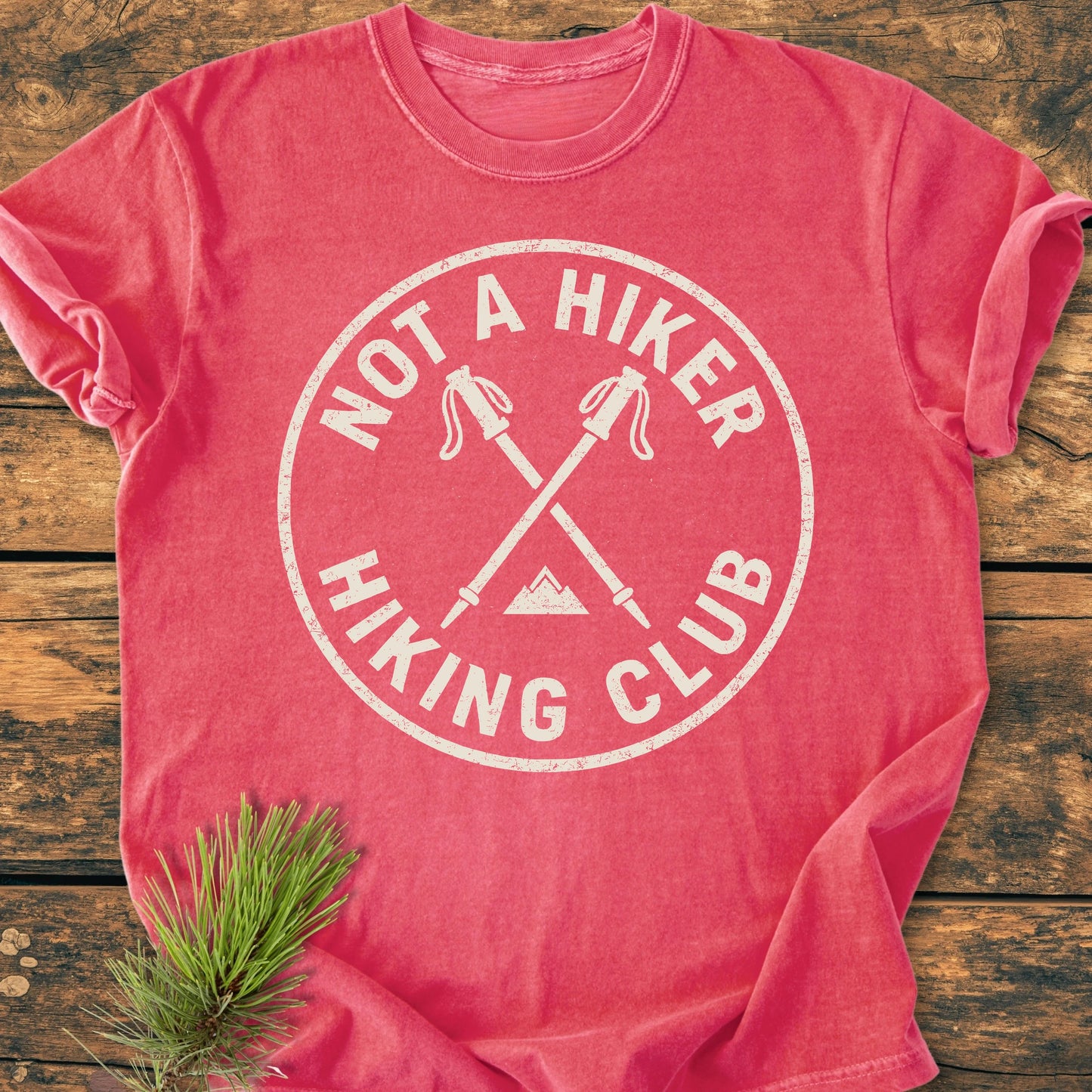 Pink-red short-sleeved T-shirt, 1426 Not A Hiker, with a circular logo featuring crossed hiking poles and a mountain, laid flat beside a pine sprig on wood.