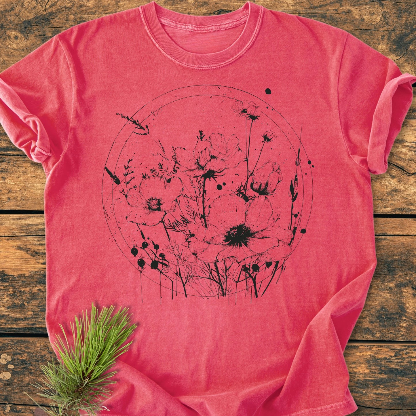 Coral-red 391 Wildflower Circle T-shirt with rolled sleeves, featuring a black wildflower graphic. Laid flat on wooden surface, with pine sprig beside it. Cozy, relaxed fit, garment-dyed fabric.