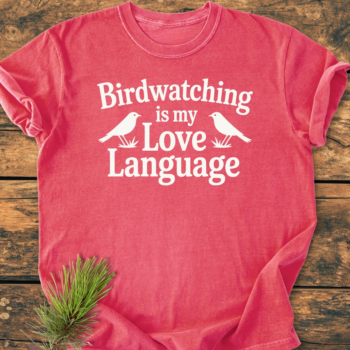 Faded-red T-shirt with “Birdwatching is my Love Language” in white, laid on wood. Features bird silhouettes and a pine sprig near hem. Relaxed unisex fit, vintage-wash for everyday wear.