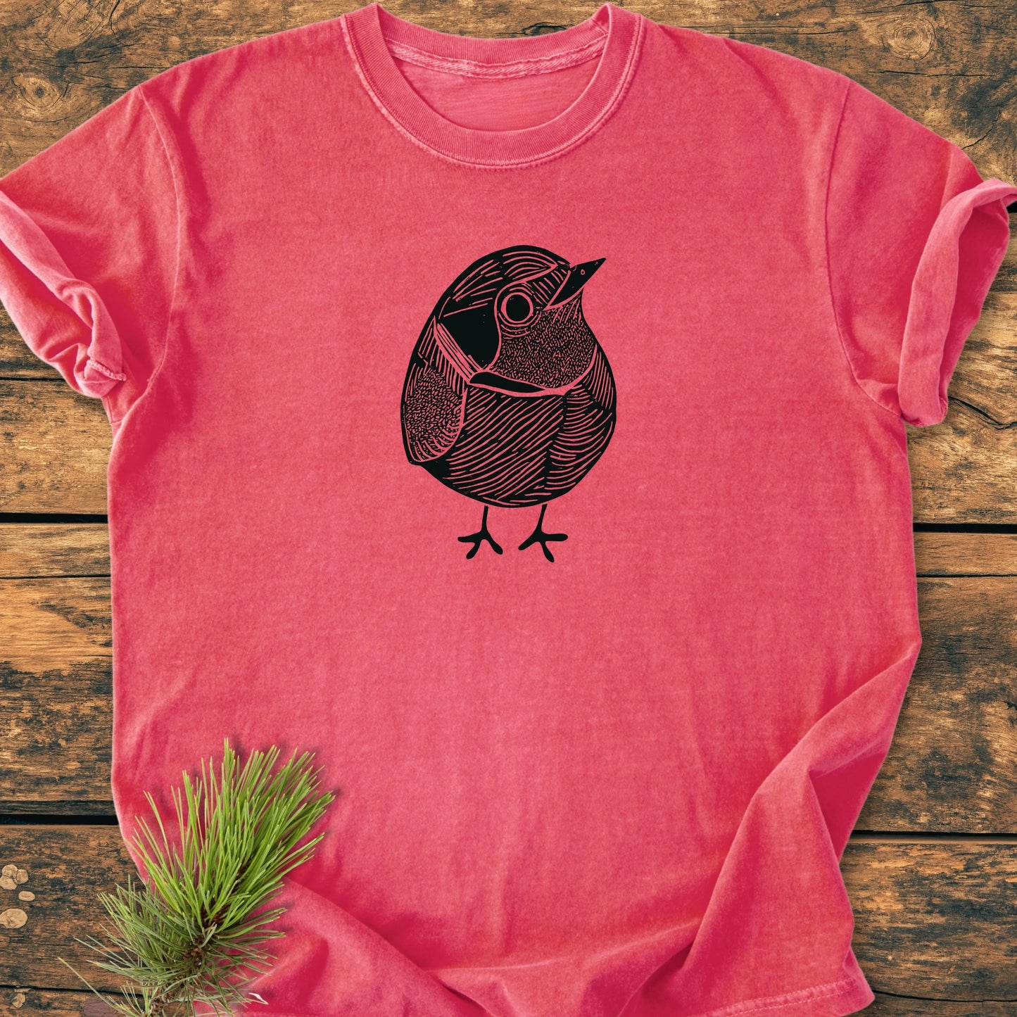 Heather-red tee with black line-art 1433 Little Robin illustration, laid flat on wooden surface; pine needles beside. Vintage wash for a soft, relaxed fit, ideal for everyday adventures.