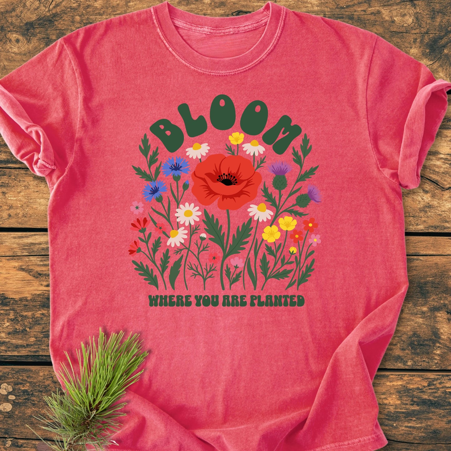 1403 Bloom Where You Are Planted Base T-shirt features a wildflower bouquet print and chunky green text, laid on wood with a pine sprig accent. Made from soft, durable ring-spun cotton.